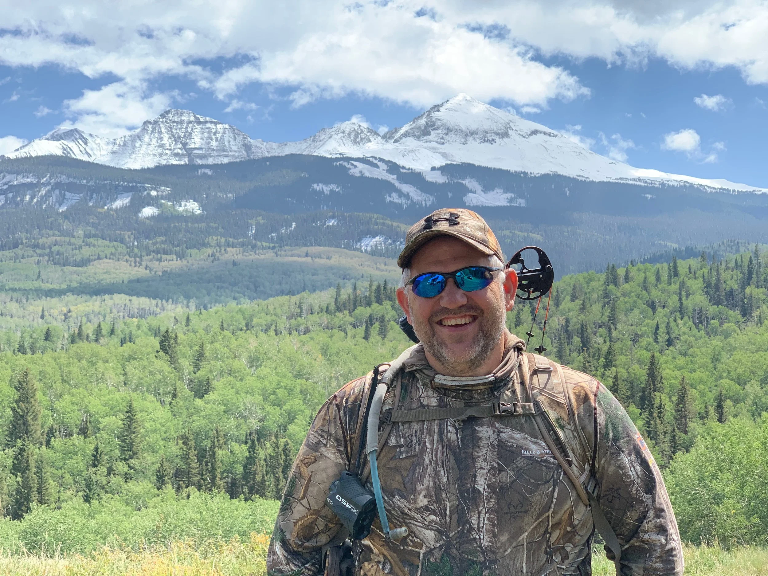 A smiling man in camouflage clothing and sunglasses standing in front of a green forest with snow-capped mountains and a blue sky in the background.