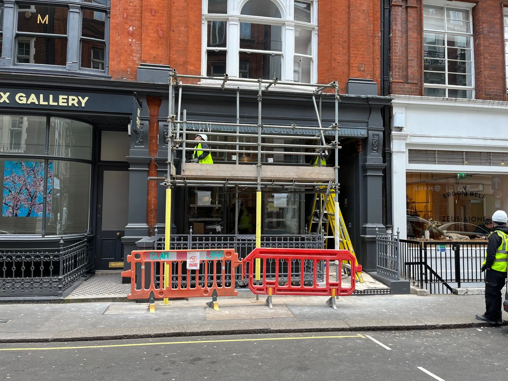 Construction workers on scaffolding working on a building facade in an urban street, with barriers in front of the storefront.