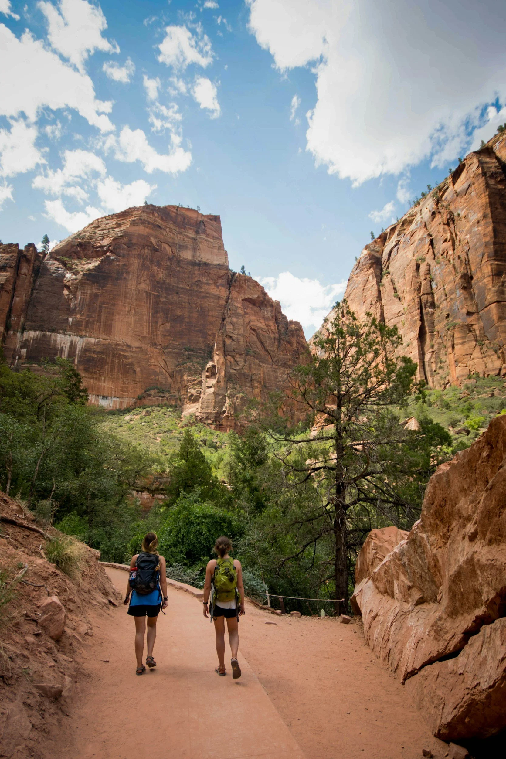 Two people hiking on a trail in a canyon surrounded by tall red rock cliffs and green trees, under a partly cloudy sky.