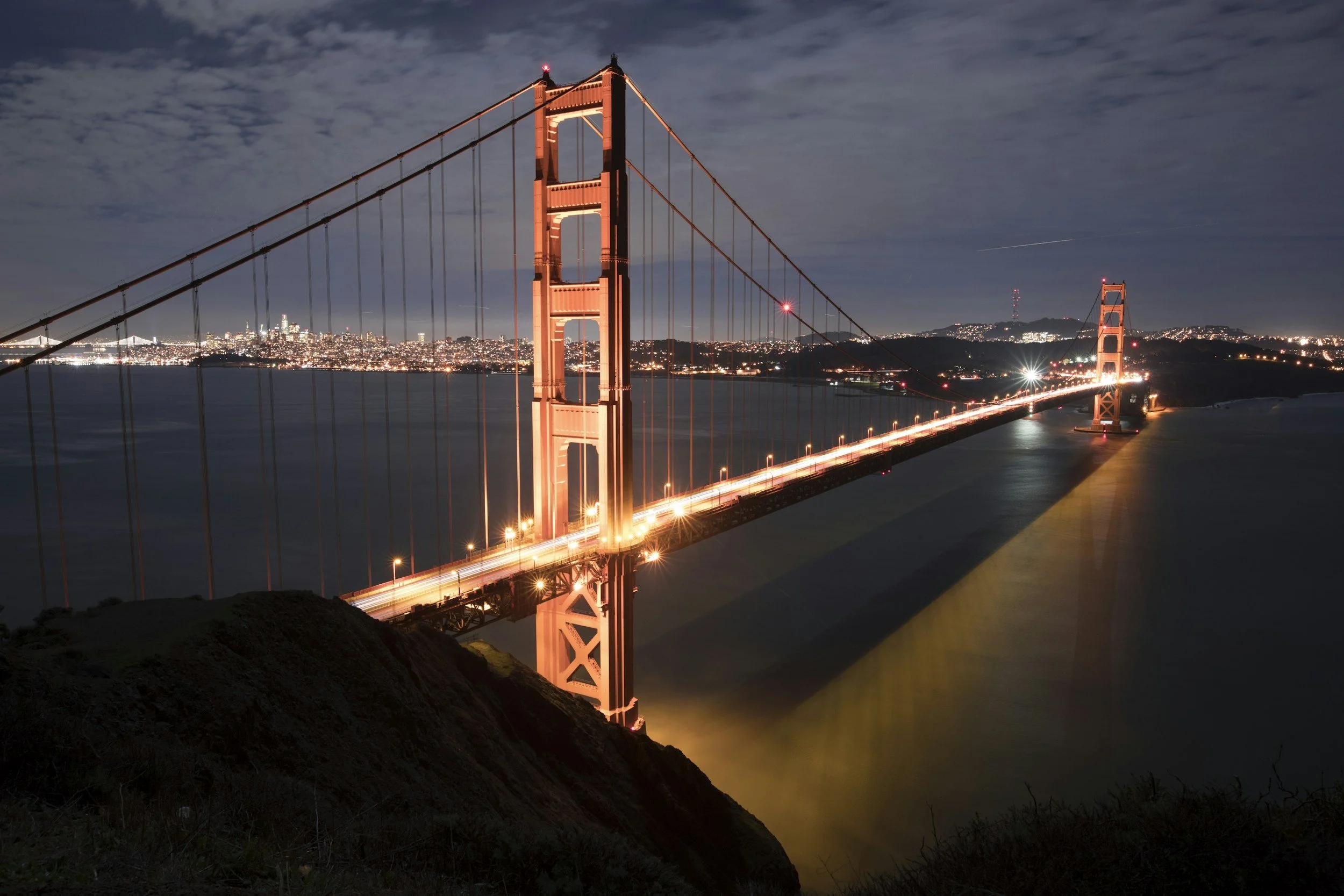 Night view of the Golden Gate Bridge in San Francisco illuminated with orange lights, with city lights in the background and a cloudy sky