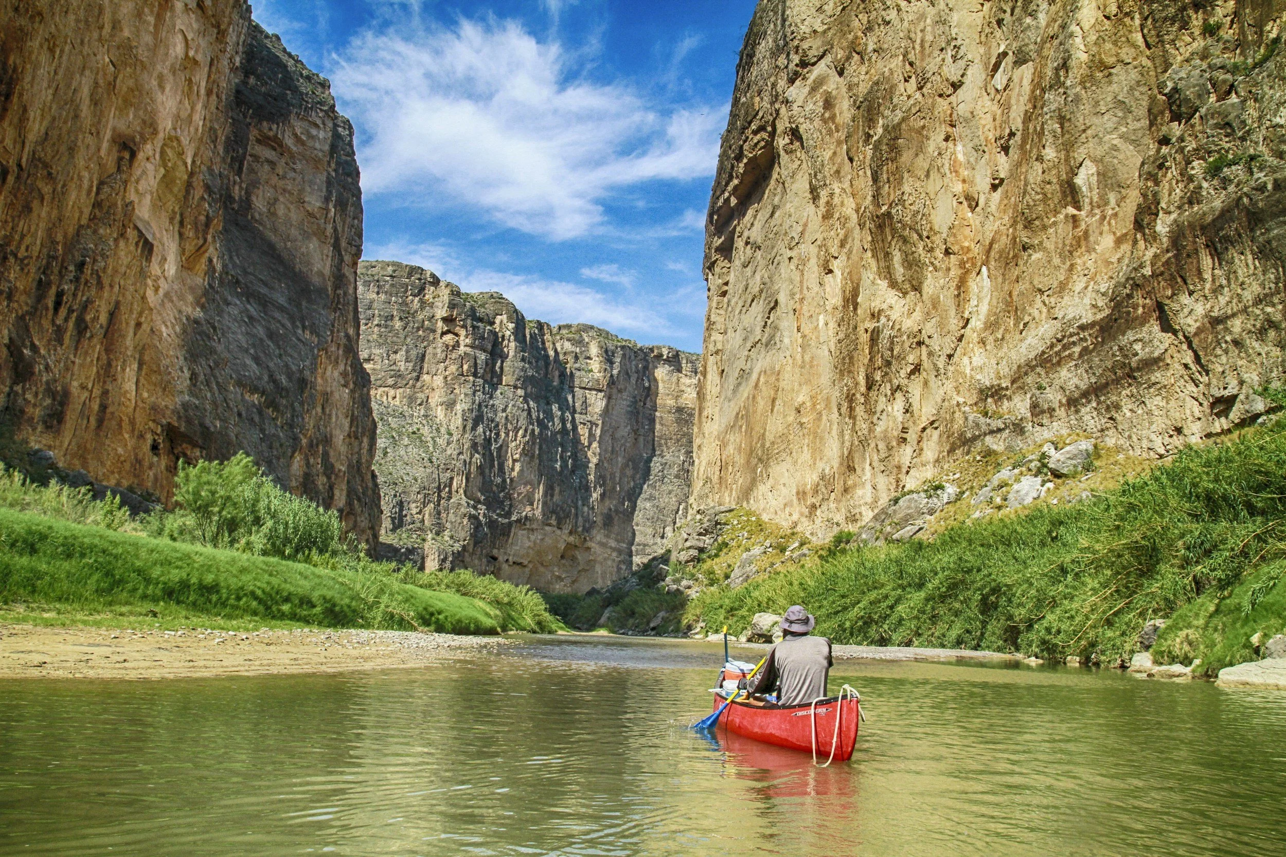 A person in a red canoe paddling on a river between towering canyon walls with green shrubs and a partly cloudy sky.