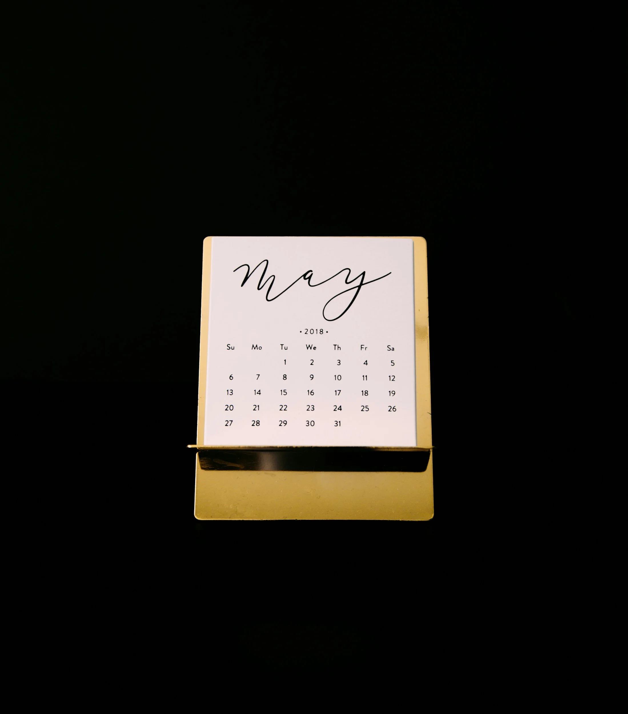 A small desktop calendar for May 2018 on a gold stand, with a black background.