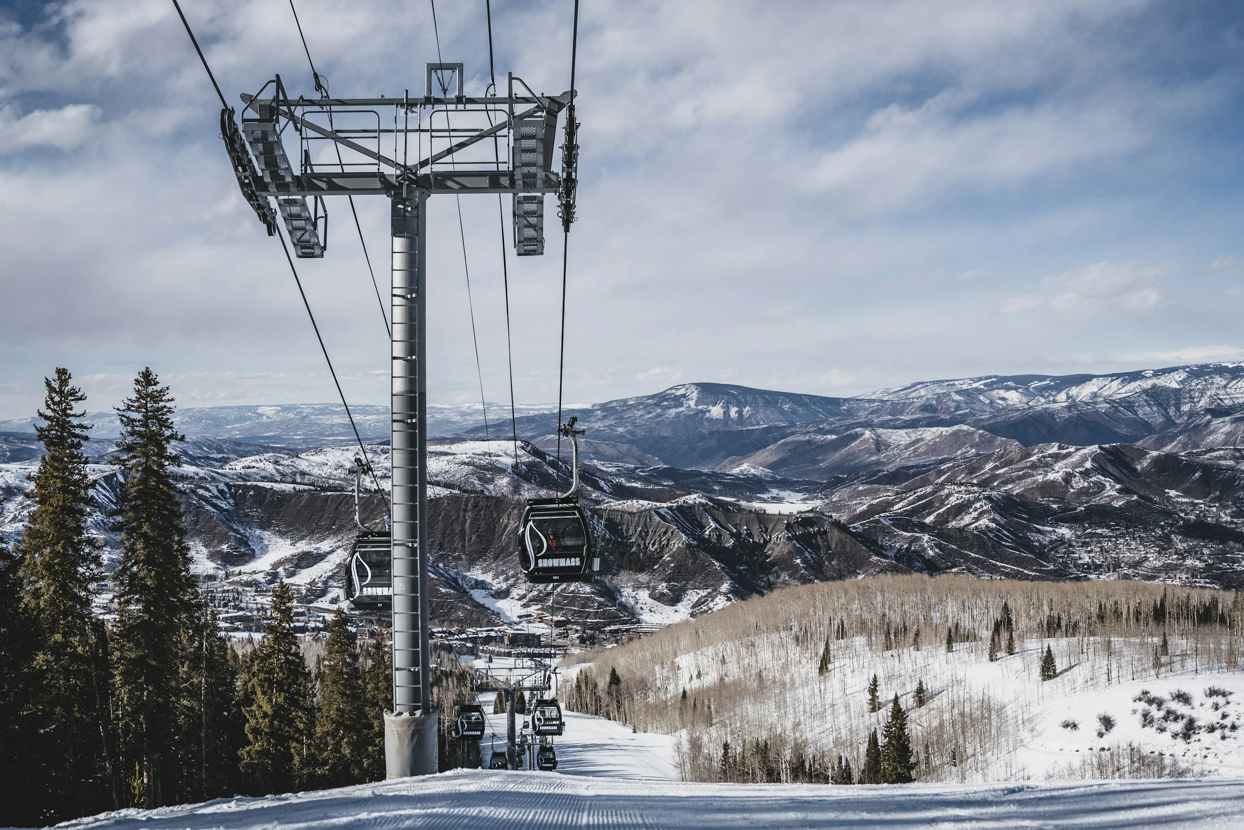 Ski lift chairs on a snowy mountain slope with a mountainous landscape in the background, trees, and partly cloudy sky.