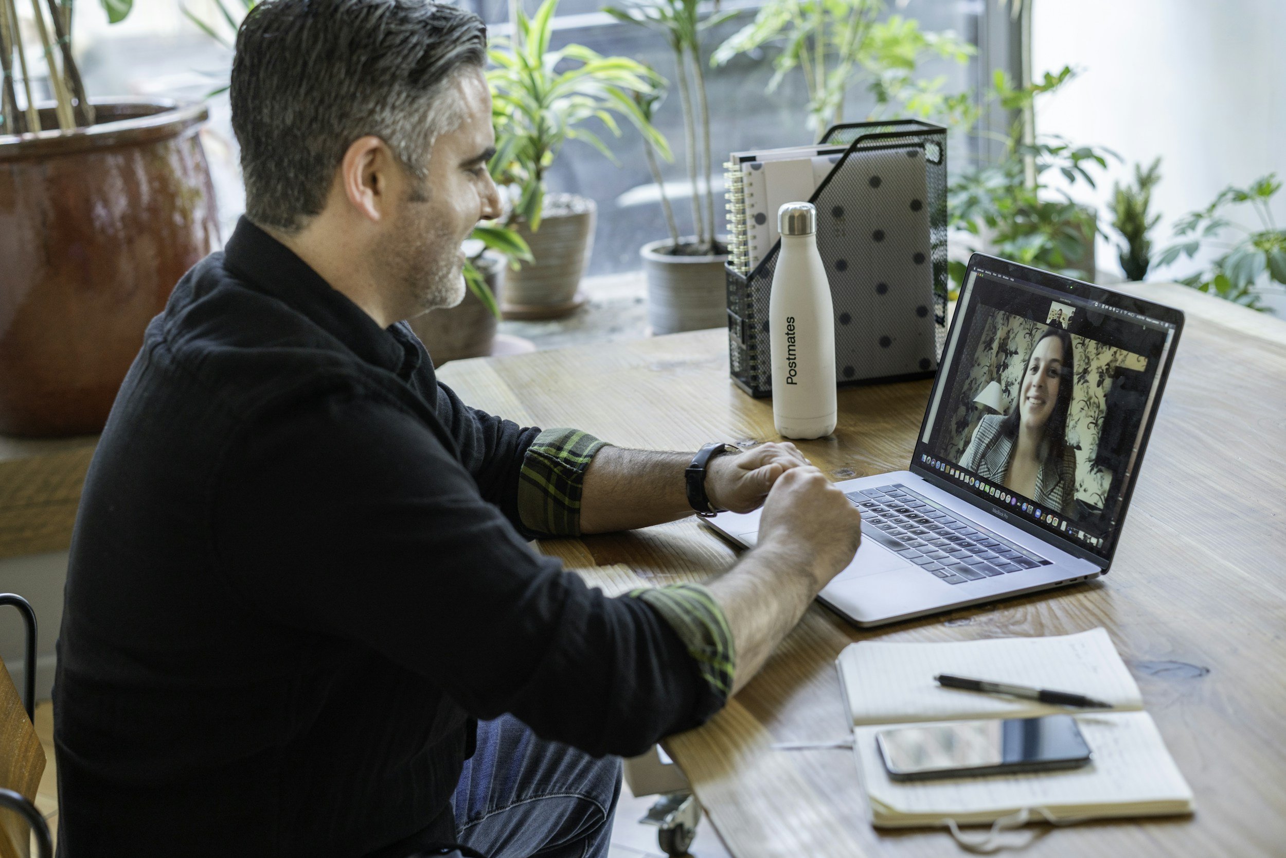 A man smiling during a video call on his laptop in a bright, plant-filled room.
