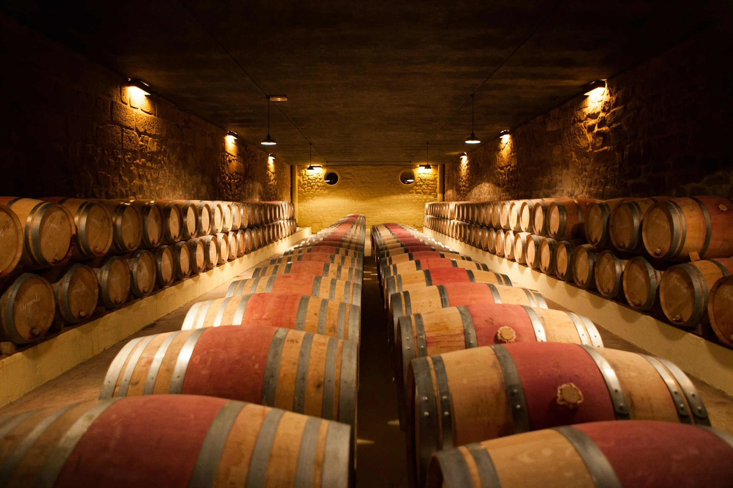 A wine cellar with wooden barrels stacked on shelves along the walls and on tables in the center, dimly lit by small ceiling lights.