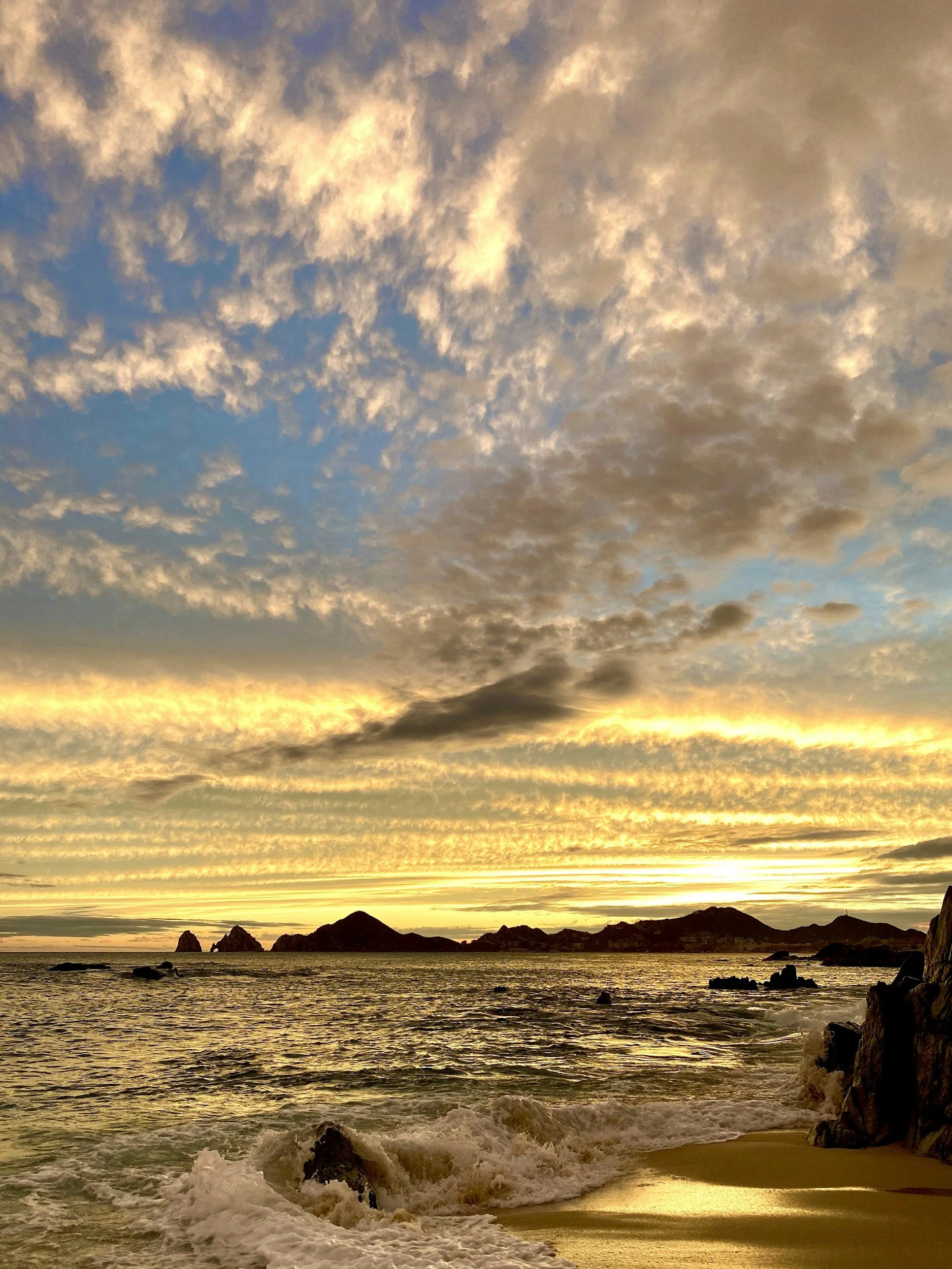 Sunset over the ocean with rocky islands, calm waves, and a partly cloudy sky.