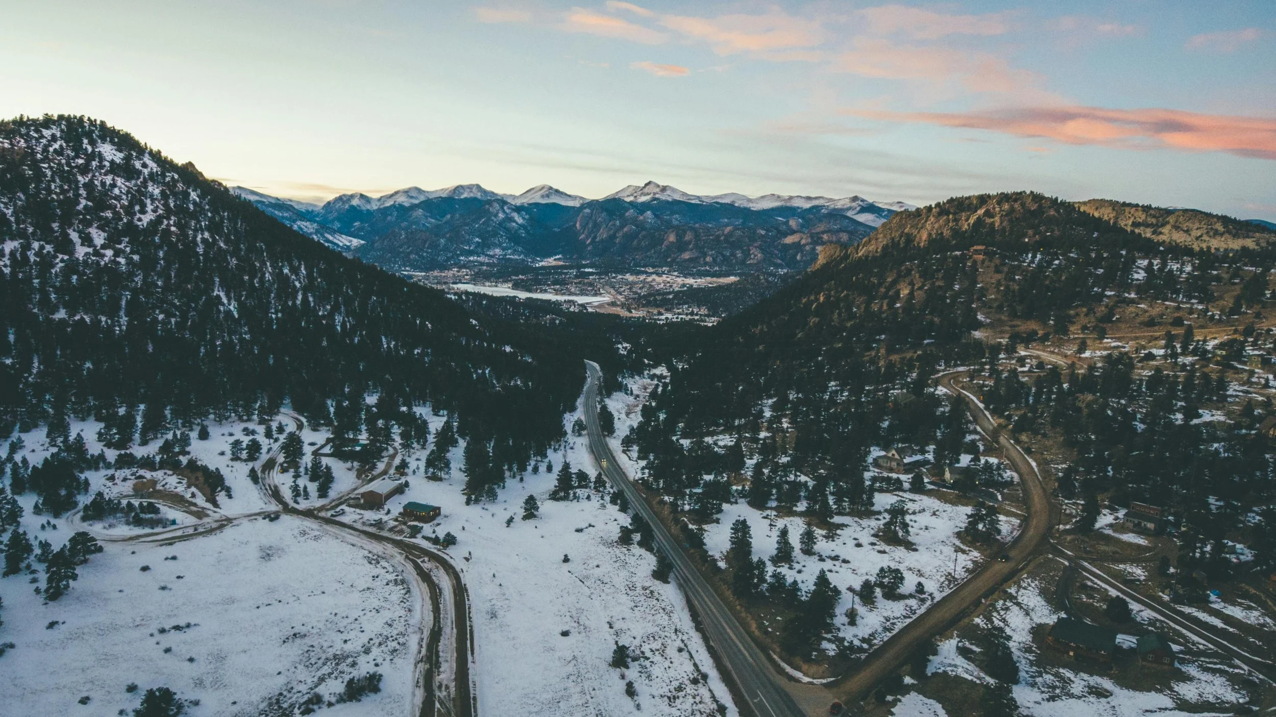 Aerial view of a snow-covered valley with winding roads, surrounding forested mountains, and a distant town under a colorful sky at sunset.