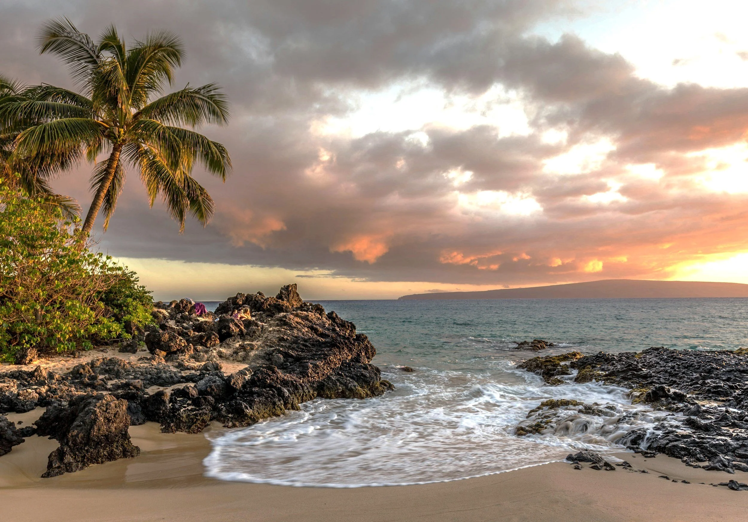 A tropical beach scene with palm trees, rocky shoreline, sand, ocean waves, and a colorful sunset sky with clouds.