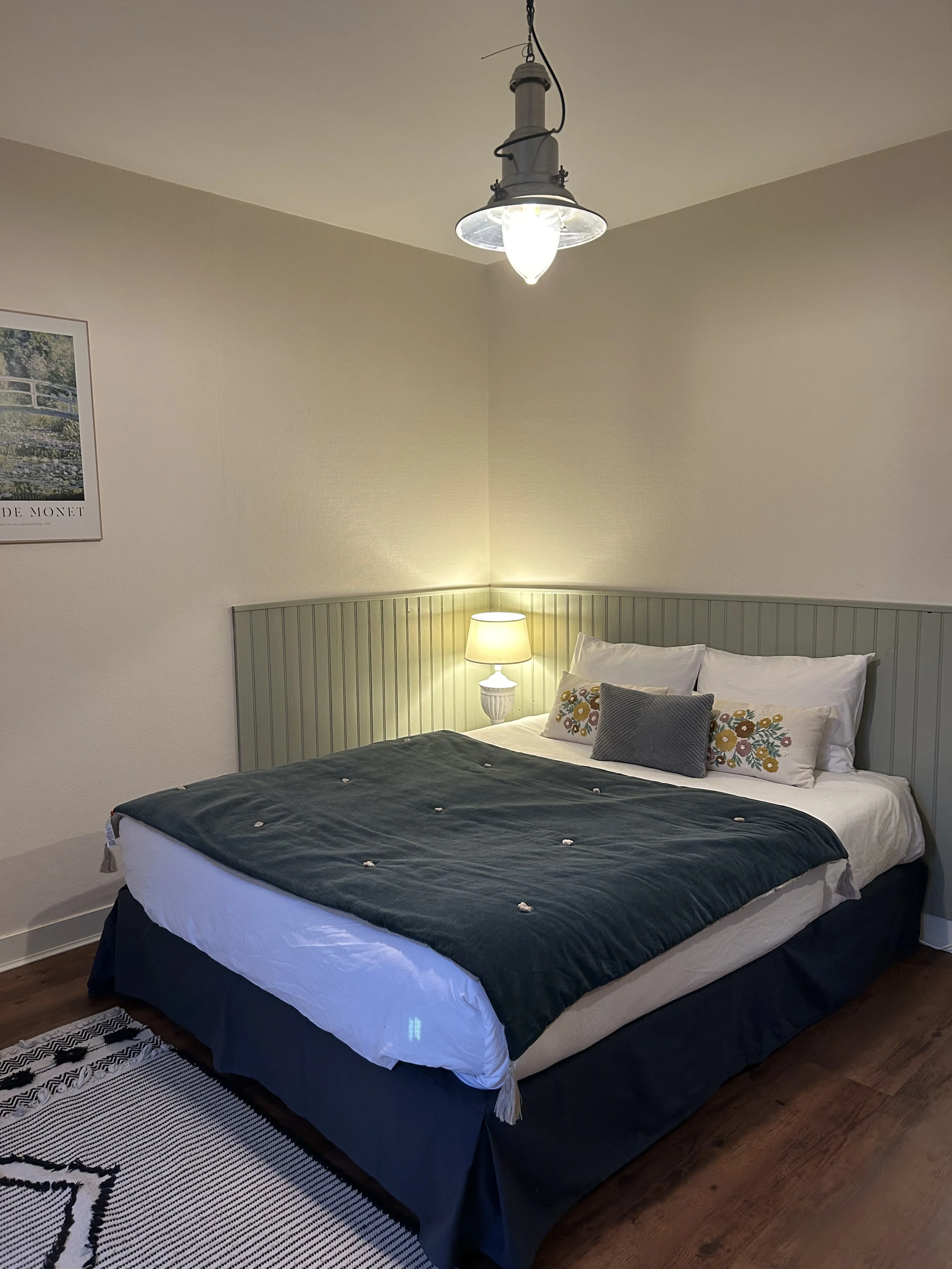 Chambre avec lit fait, draps blancs, couverture verte, coussins décoratifs, lampe de table, peinture, et luminaire suspendu au plafond.