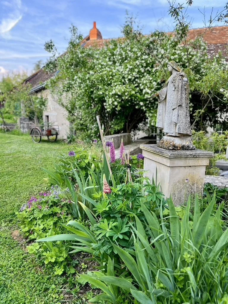 Statue en pierre d'une figure religieuse dans un jardin avec des fleurs colorées et un buisson fleuri en arrière-plan.