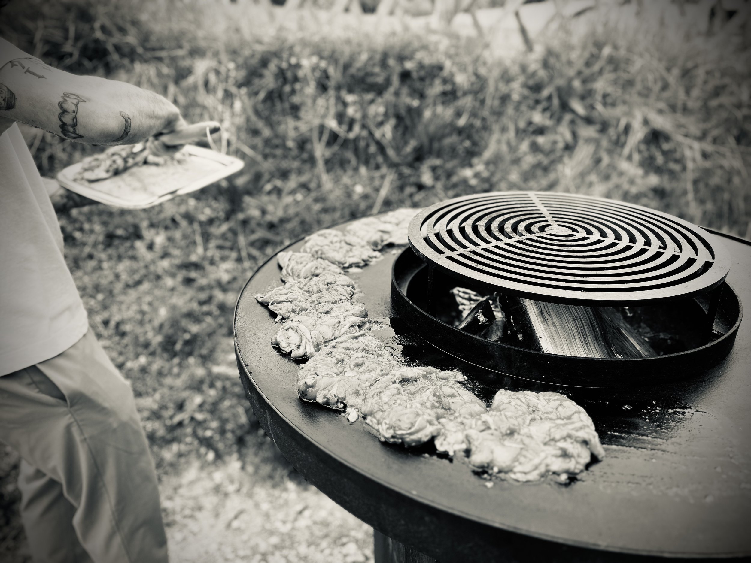 Personne faisant cuire des morceaux de viande sur un barbecue en plein air, en noir et blanc.