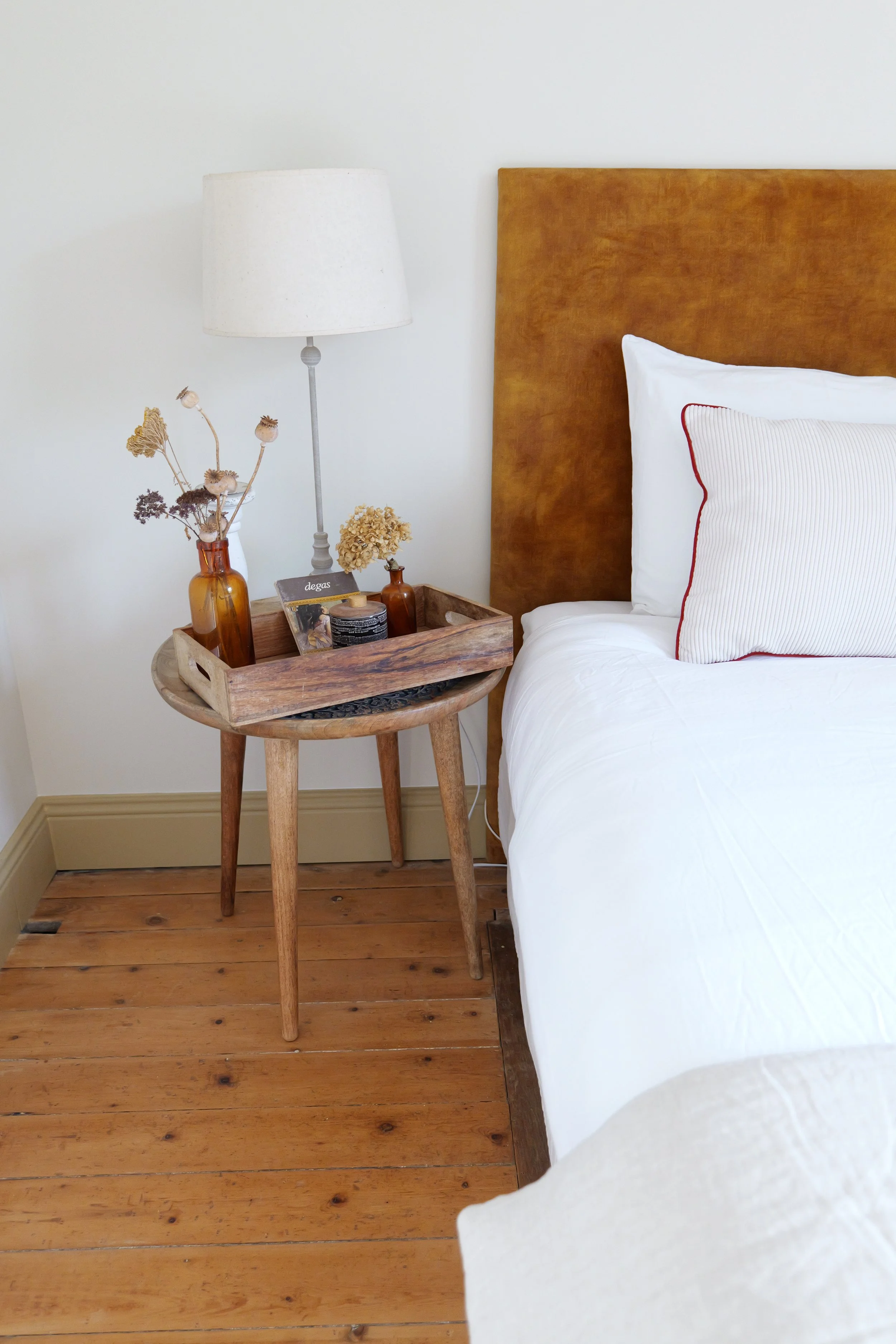 Coin de chambre avec une table de chevet en bois, une lampe à abat-jour blanc, des bouteilles en verre avec des fleurs séchées, et un lit avec une tête de lit en cuir brun, des draps blancs et un coussin.”}