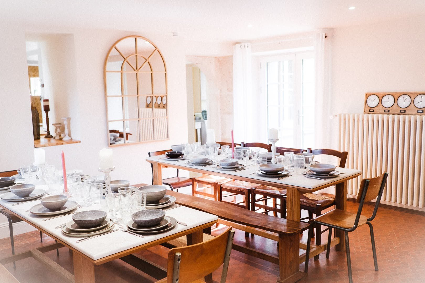 Salle à manger lumineuse avec une grande table en bois, vaisselle blanche, verres, bougies, et un miroir déco sur le mur