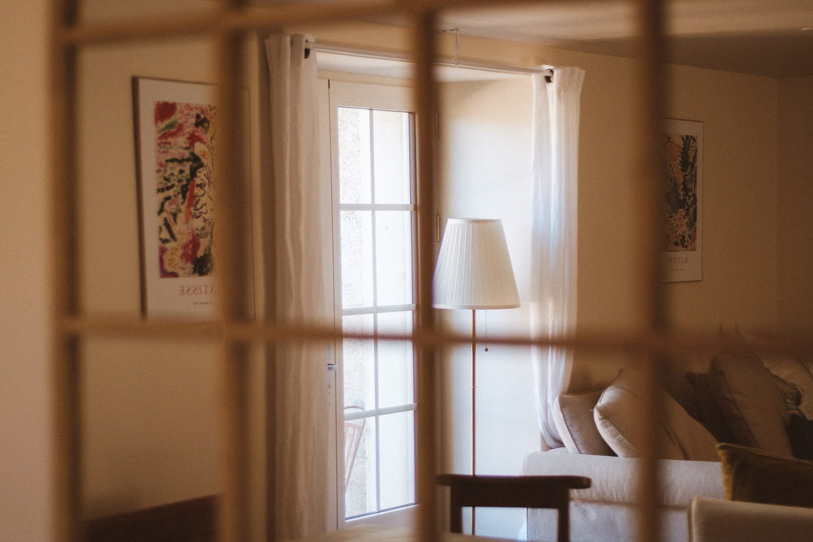 Intérieur d'un salon vu à travers une grille, comprenant un canapé beige, une lampe de lecture, des rideaux blancs, une porte-fenêtre, et des œuvres d'art modernes aux murs.