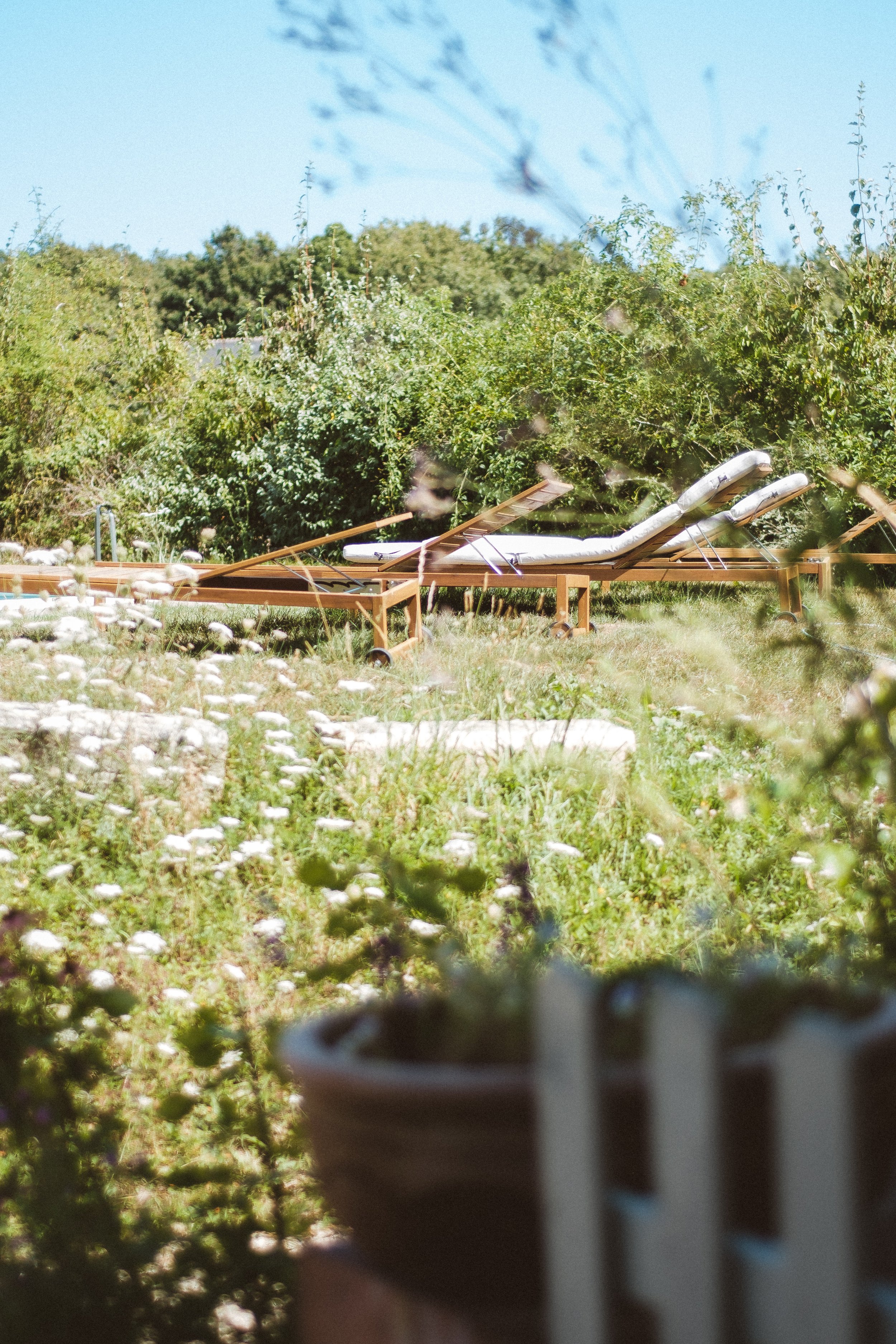 Deux chiliennes longues en blanc sur un lit bain de soleil en bois, dans un jardin verdoyant en plein air, avec une végétation luxuriante en arrière-plan et un ciel bleu clair.