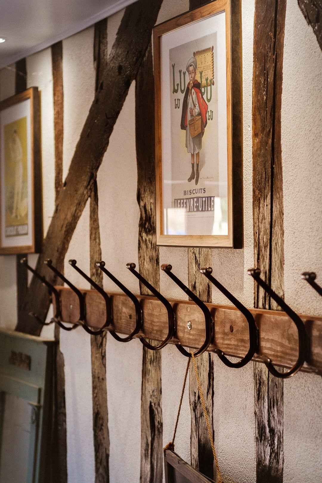 Murs en bois apparents avec une rangée de six crochets en métal, une affiche ancienne avec un homme en costume et un chapeau, des cadres avec des illustrations, et une échelle en bois suspendue avec une corde.