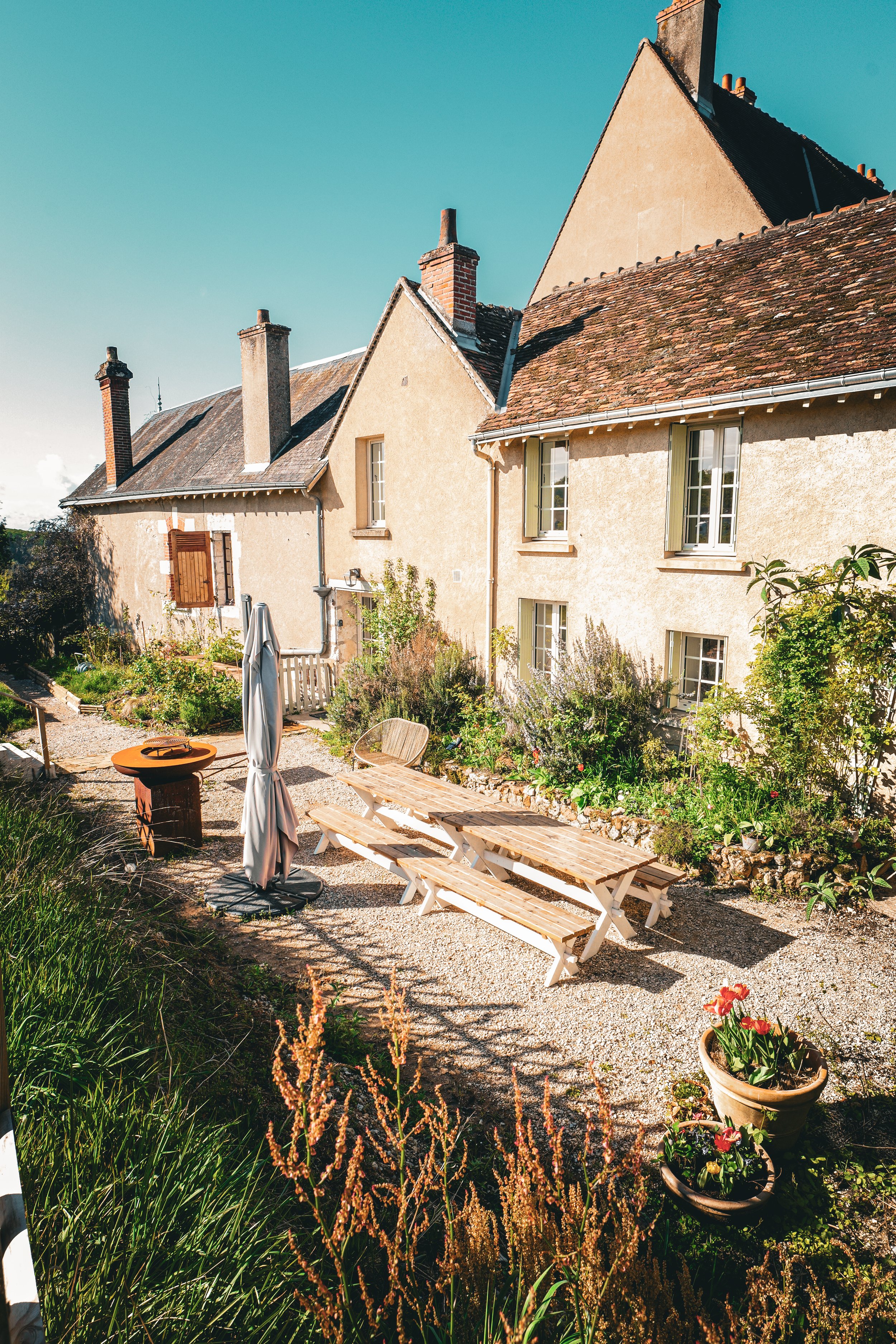 Cour arrière d'une maison ancienne avec jardin, table en bois, parasol plié, fleurs en pot et végétation verdoyante sous un ciel clair.
