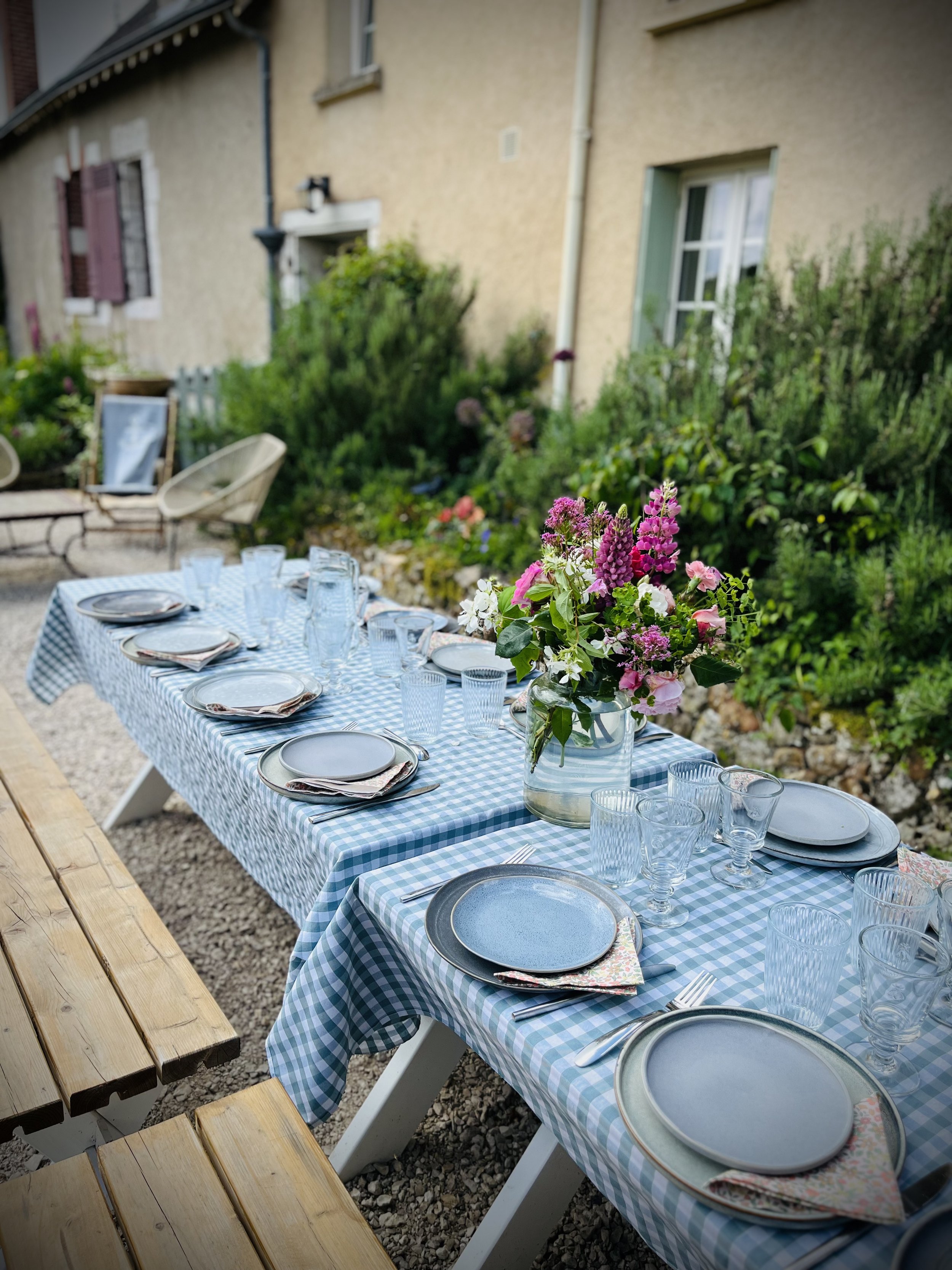 Table dressée pour un repas en extérieur avec une nappe à carreaux bleus et blancs, des assiettes, des verres, des couverts, et un bouquet de fleurs colorées en vase, dans un jardin ensoleillé.
