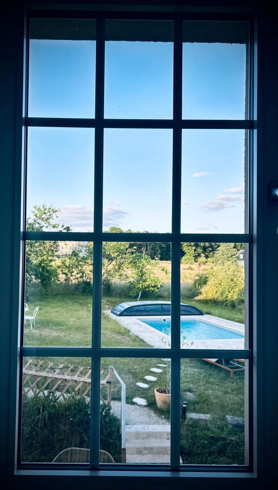 Vue à travers une fenêtre en verre divisée en neuf sections, montrant un jardin avec une piscine, des chaises, et des arbres verdoyants sous un ciel bleu avec quelques nuages.