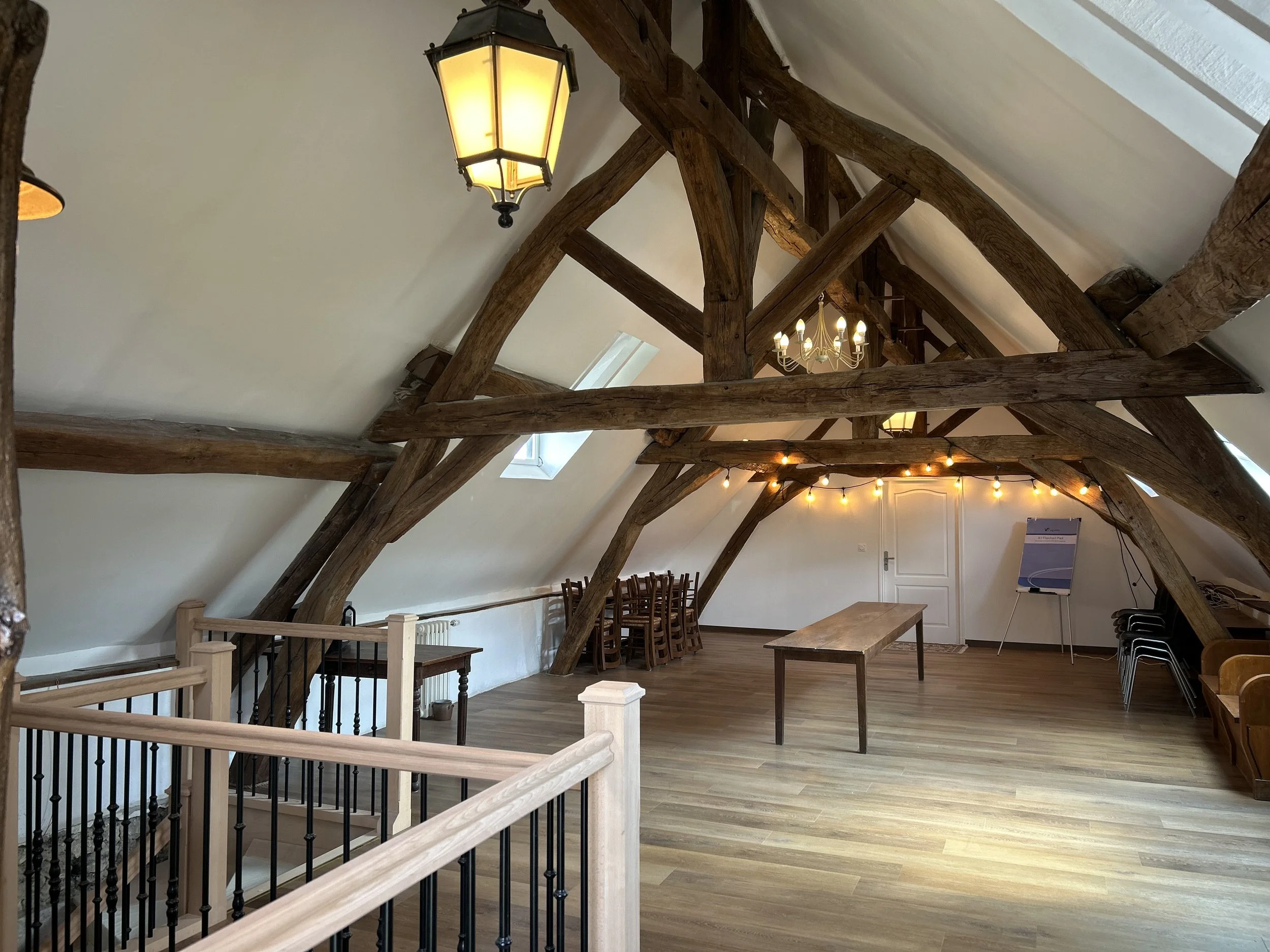 Intérieur d'une grande salle mansardée avec poutres en bois apparentes, un lustre blanc, lampes suspendues, une table en bois, et une chaise pliante. Mur blanc avec une porte à l'arrière et un tableau blanc sur un chevalet.