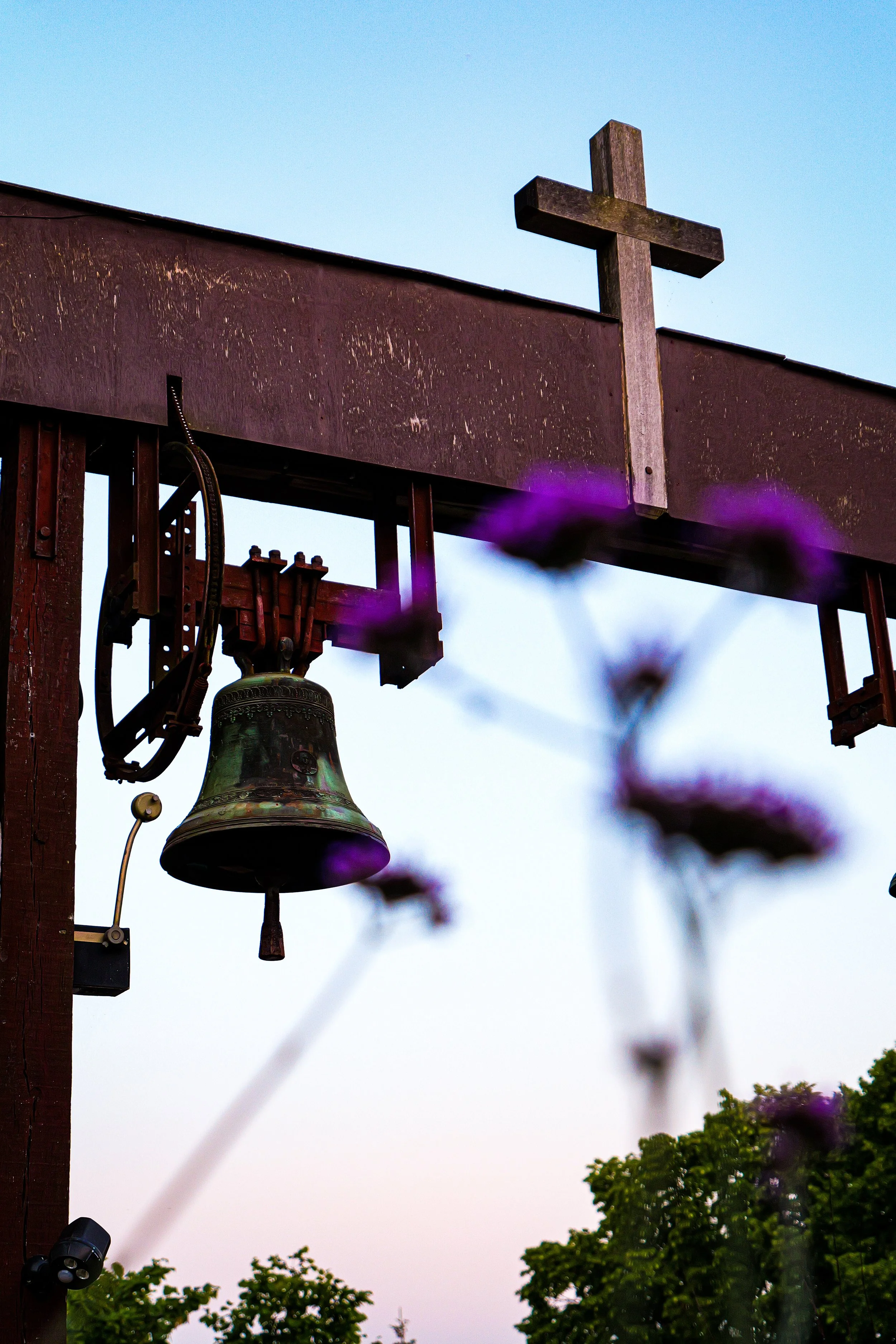 Une cloche suspendue sous une croix en bois, avec des fleurs violettes floues au premier plan et un ciel clair en arrière-plan.