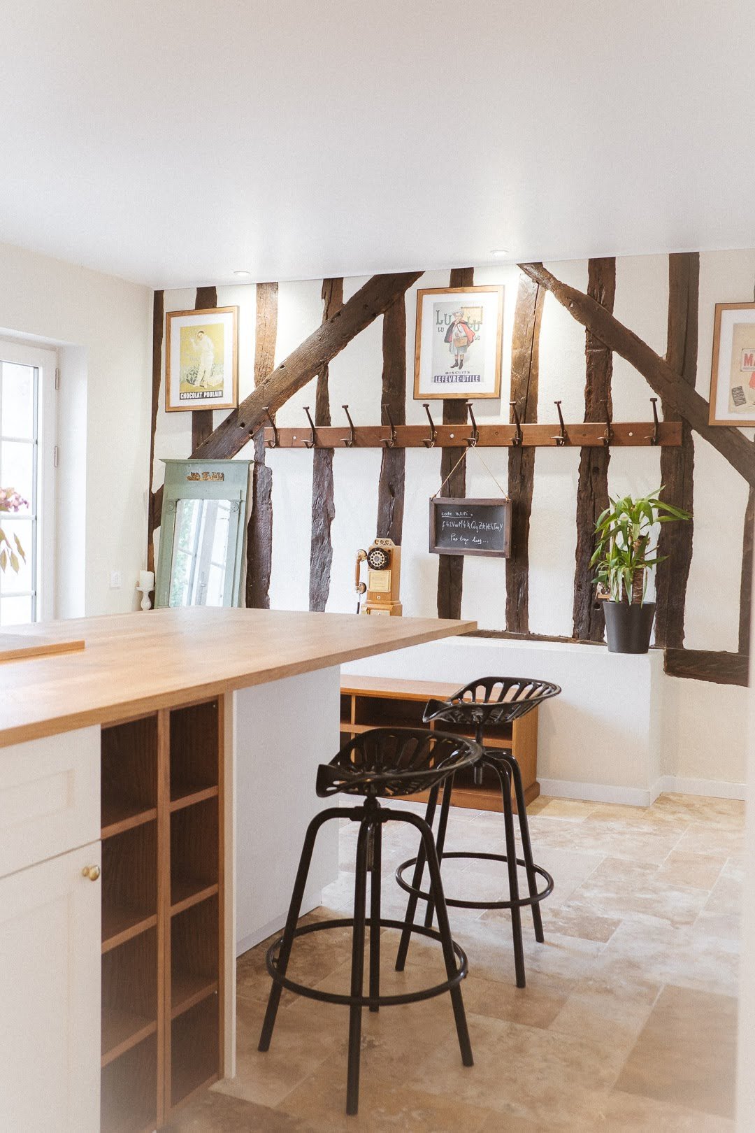 Intérieur d'une cuisine avec un comptoir en bois, deux chaises noires en métal, un mur avec des poutres en bois apparentes, un miroir ancien et des affiches encadrées.
