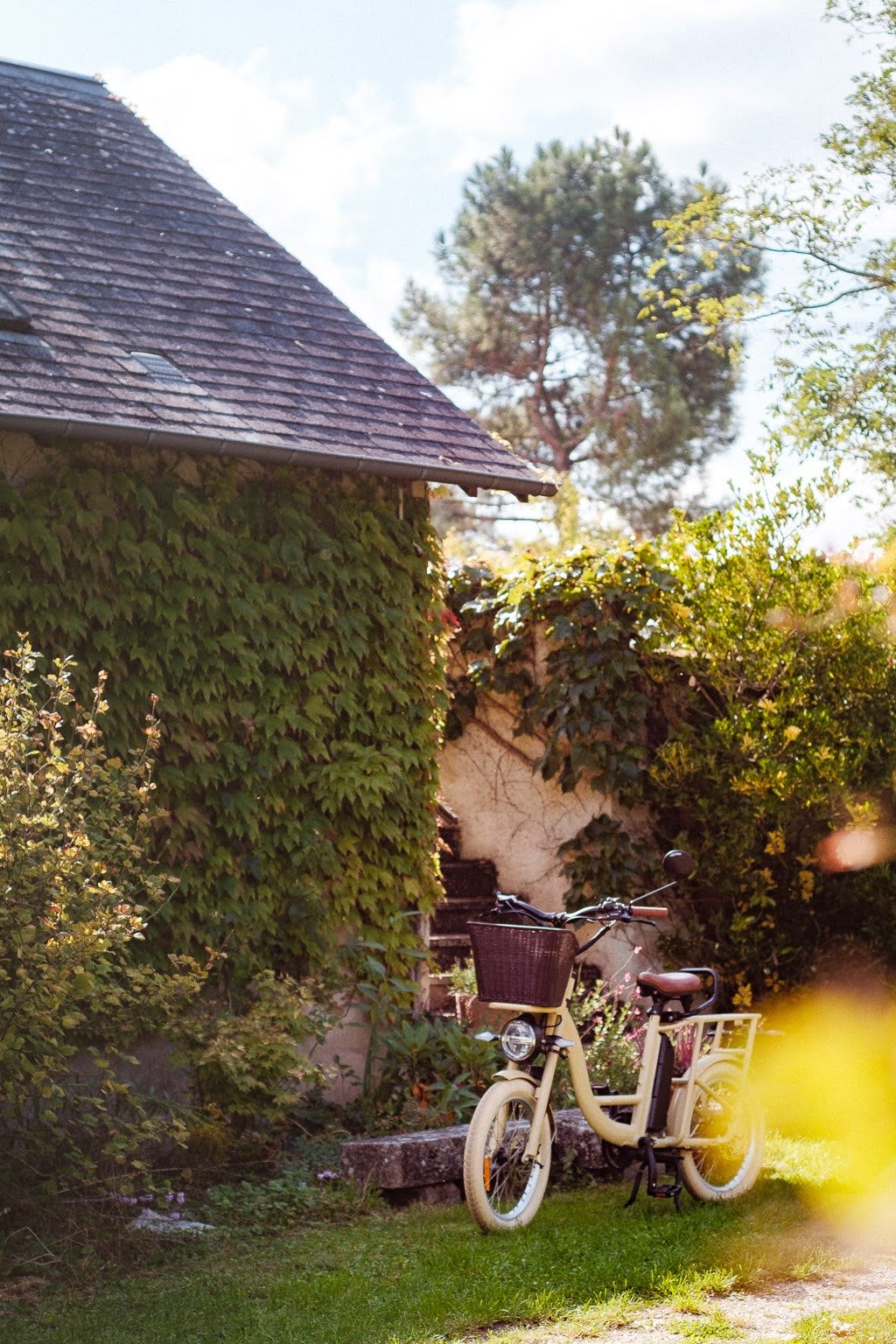 Vélotaxi beige stationné dans un jardin avec des plantes leafy, à côté d'une maison partiellement couverte de végétation, par une journée ensoleillée.