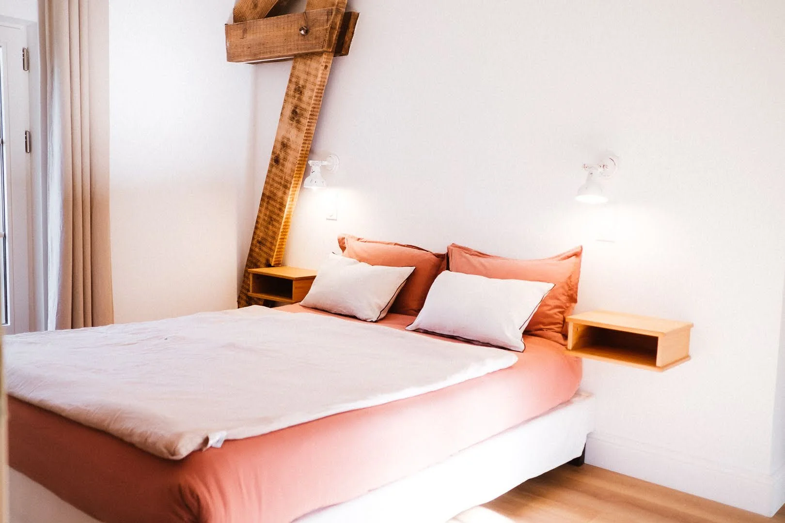 Chambre avec lit à draps rose, deux oreillers blancs, table de nuit en bois et lumières murales