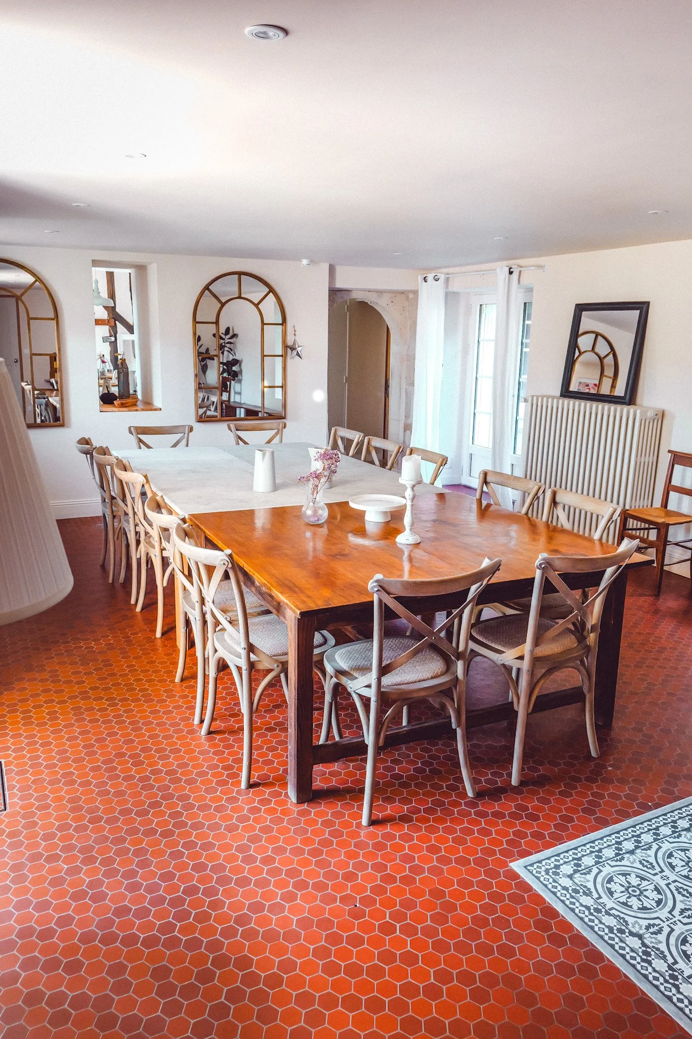 Salle à manger avec une grande table en bois, entourée de chaises en bois, décorée de vases, chandelles et assiettes, avec des miroirs à cadre doré et fenêtres avec rideaux blancs.