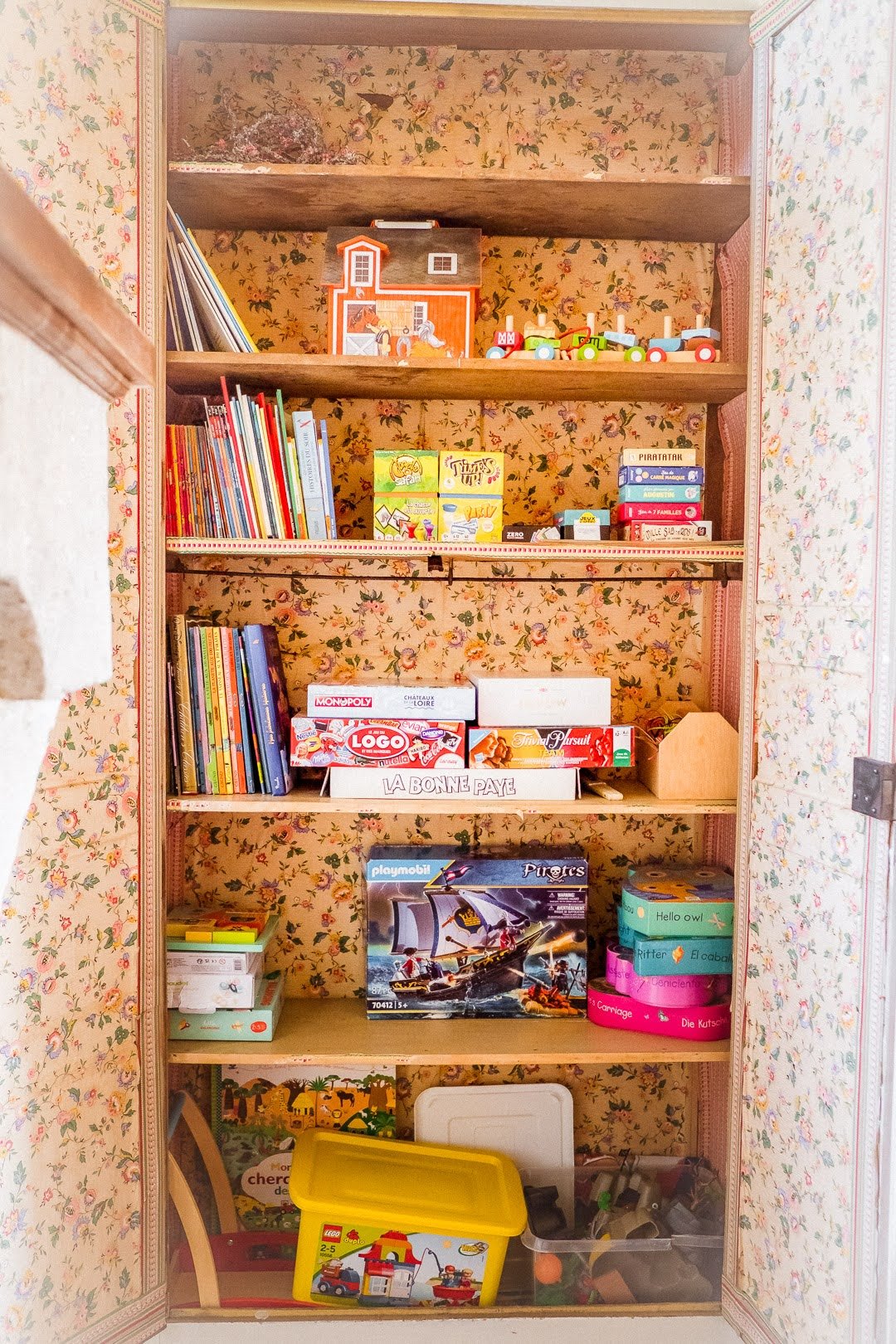 Une étagère en bois remplie de livres, jeux de société, jouets en bois, un puzzle LEGO Pirates, et autres jouets pour enfants, avec un fond de papier peint floral