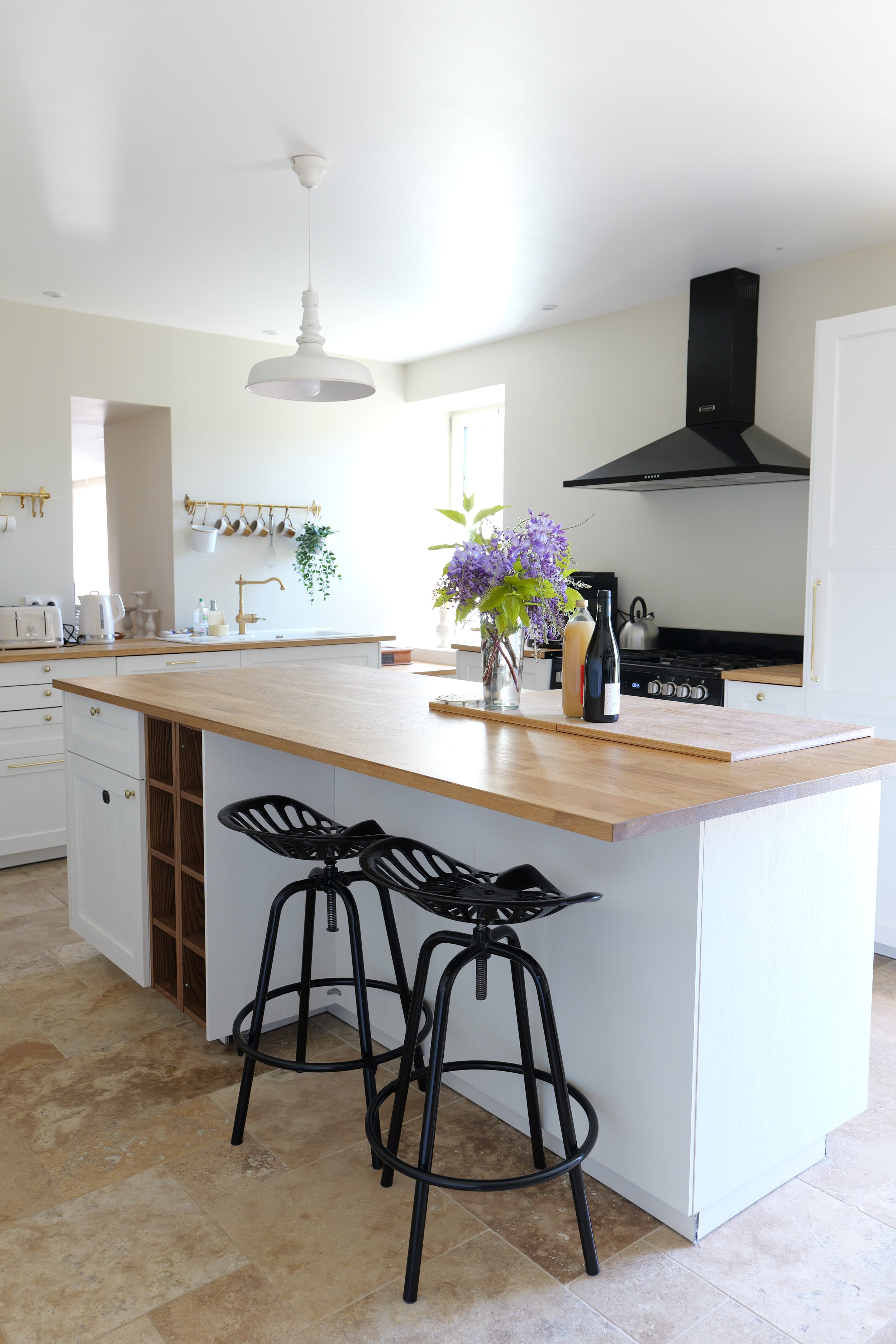 Cuisine moderne avec îlot central en bois clair, deux tabourets noirs en métal, bouquet de fleurs violettes, appareils électroménagers blanc, plinthe en bois, murs blancs, fenêtre lumineuse.