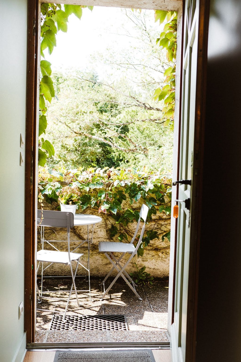 Petite terrasse en pierre avec deux chaises en métal blanc, une petite table avec un pot de fleurs, entourée de végétation verte, vue depuis une porte entrouverte.