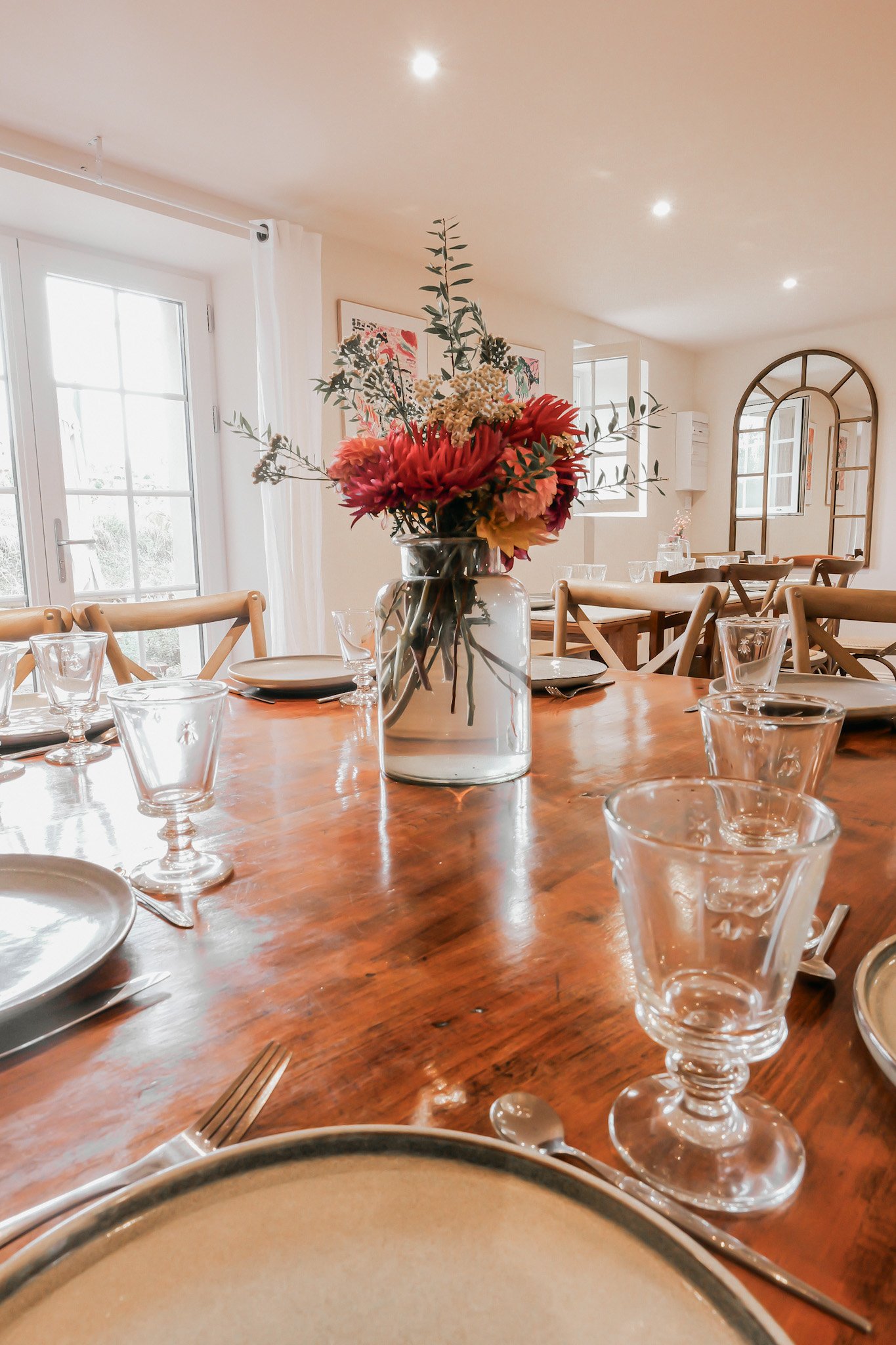 Table de salle à manger décorée avec un vase rempli de fleurs colorées, entourée de vaisselle en verre et de coupelles, dans une salle lumineuse avec fenêtres et miroirs.