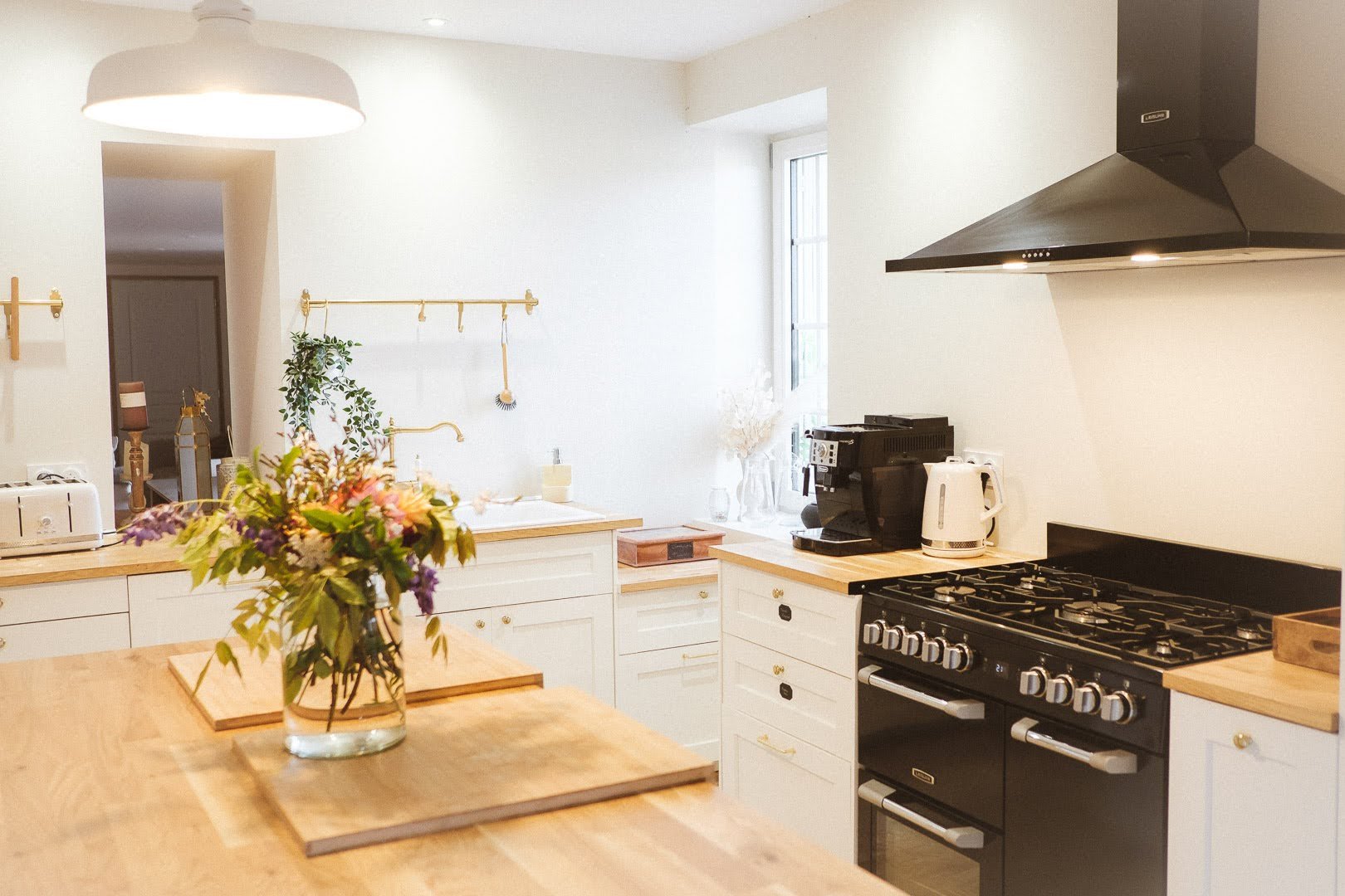Cuisine lumineuse avec plan de travail en bois, fleurs en vase, cuisinière à gaz, appareils électroménagers et fenêtres laissant entrer la lumière naturelle.