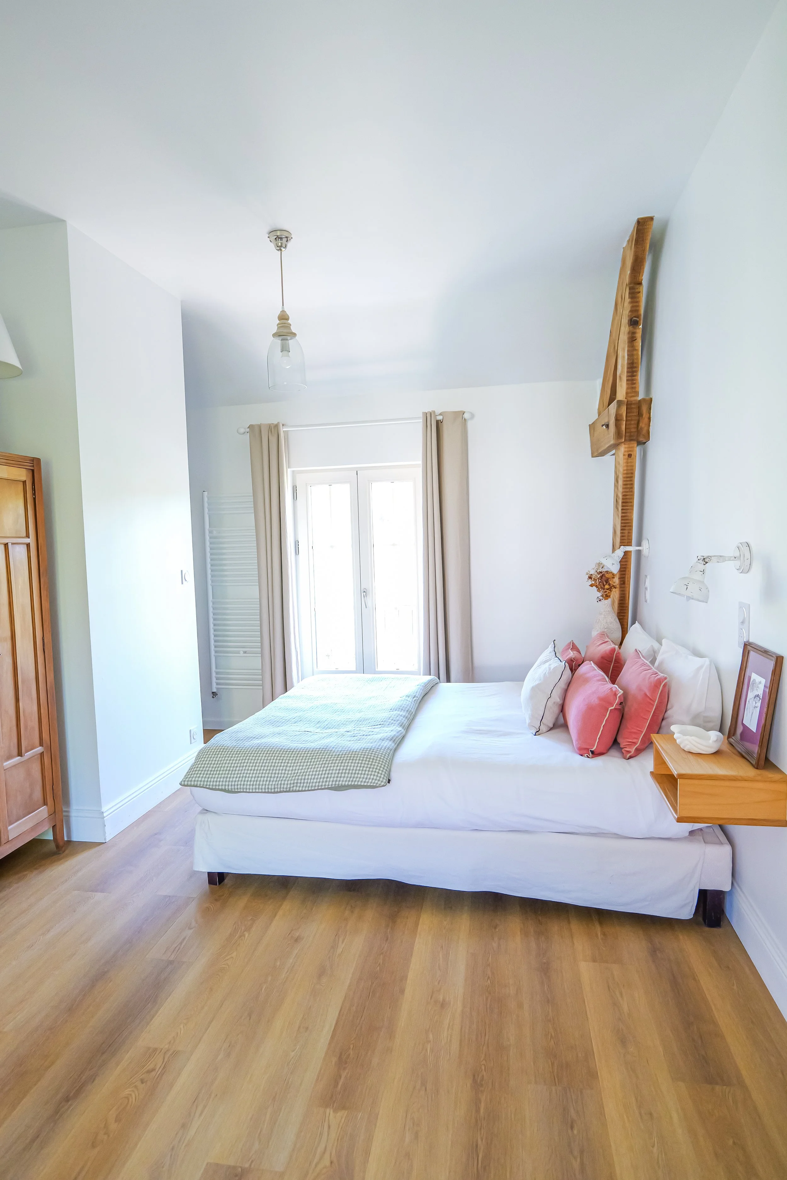 Chambre lumineuse avec lit fait, coussins roses et blanc, fenêtre avec rideaux beige, mobilier en bois, parquet en bois clair, mur blanc, lampe suspendue au plafond, décoration simple et épurée.