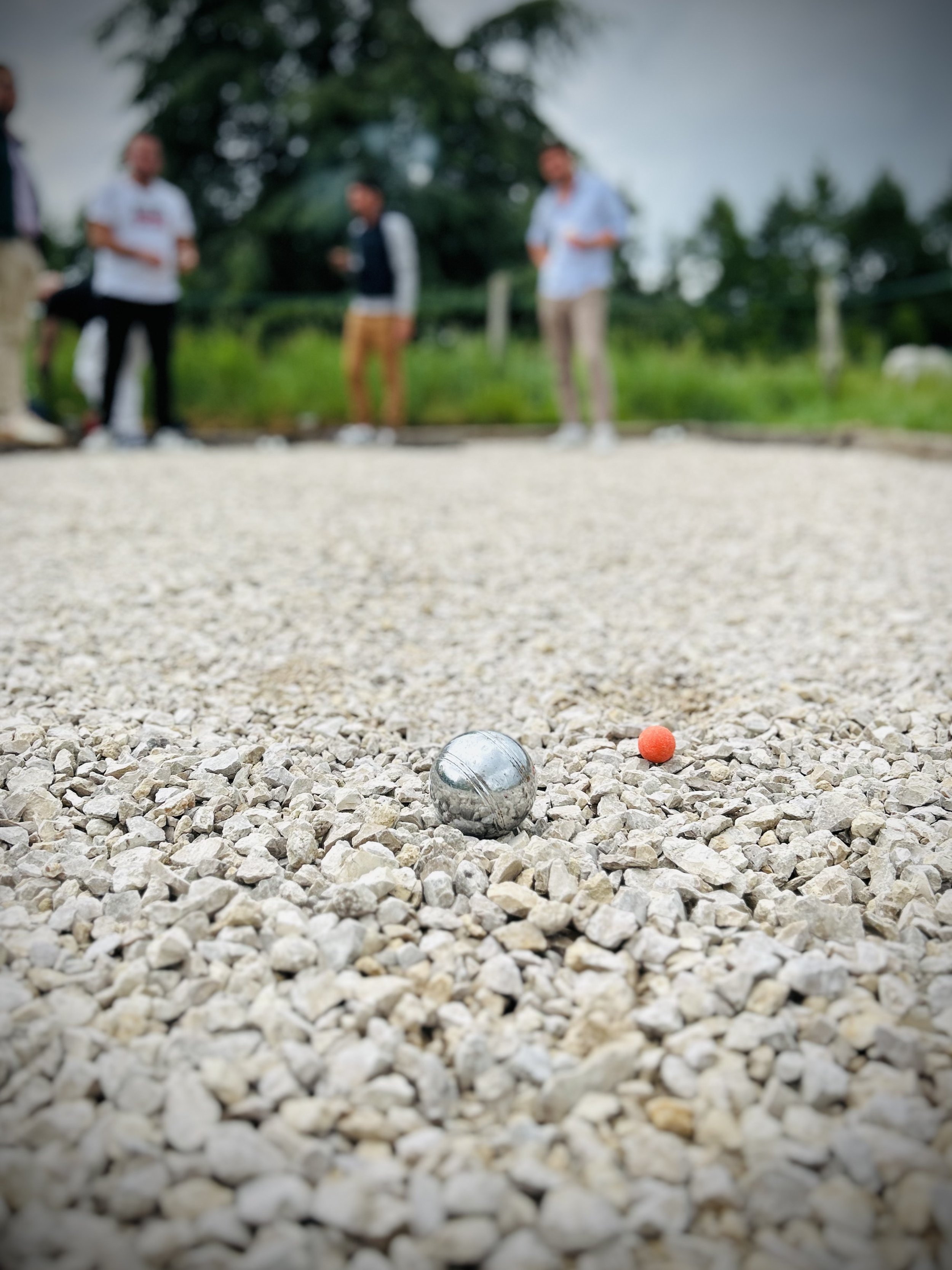 Gros plan sur une boule de pétanque en métal et une petite boule orange de pétanque sur un terrain en gravier, avec des personnes floues en arrière-plan.