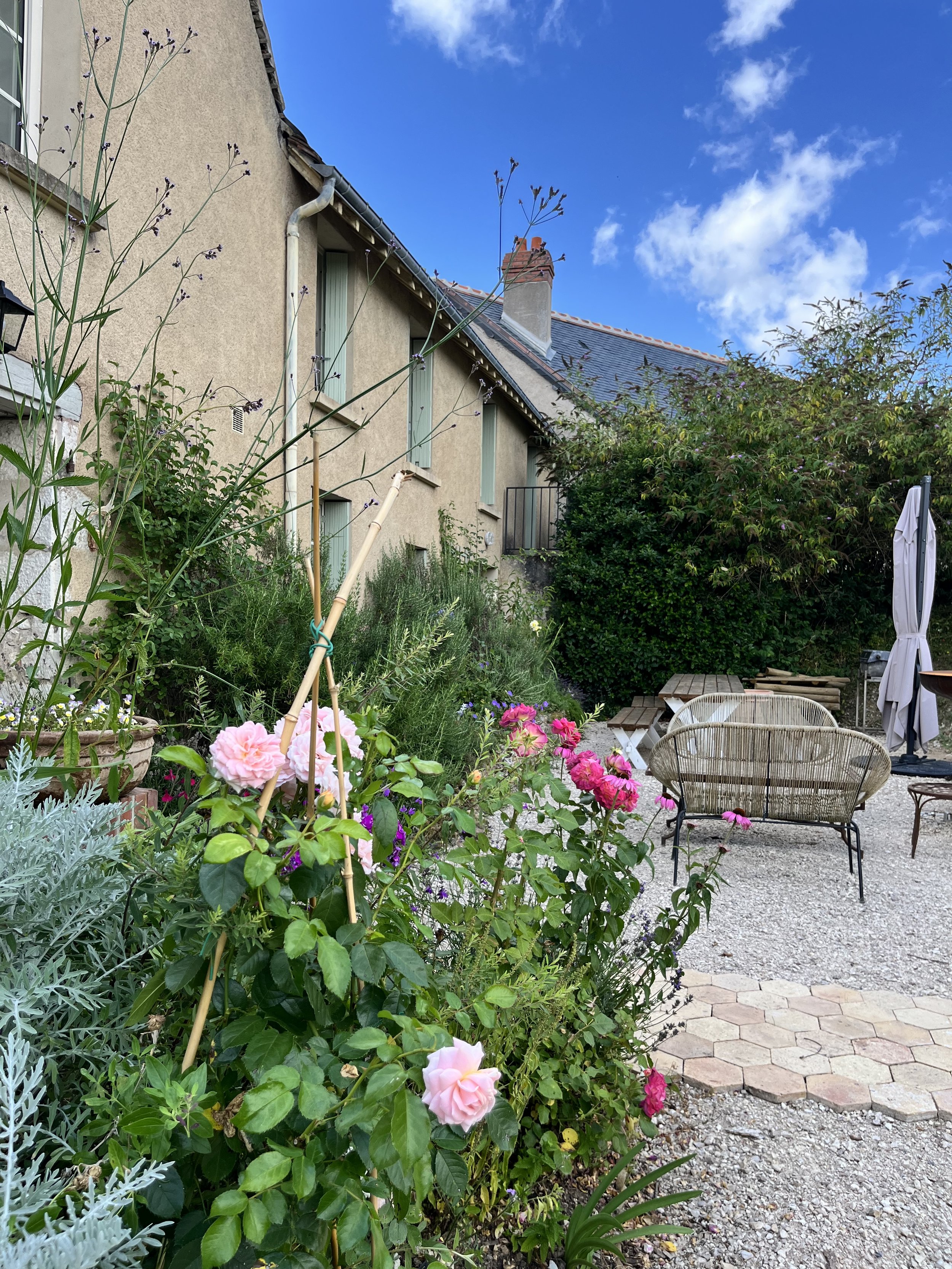 Jardin avec des fleurs roses, des meubles en rotin, un parasol fermé, et une maison ancienne en arrière-plan sous un ciel bleu avec quelques nuages.