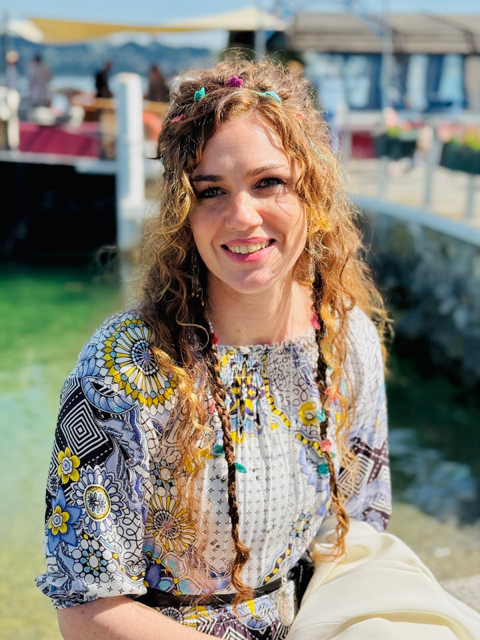 Jeune femme souriante avec des cheveux bouclés et une coiffure colorée, portant une robe à motifs floraux, devant un environnement marin avec des bateaux et une jetée. Intervenante retraite de yoga au domaine de la Chambaudière, gîte de groupe