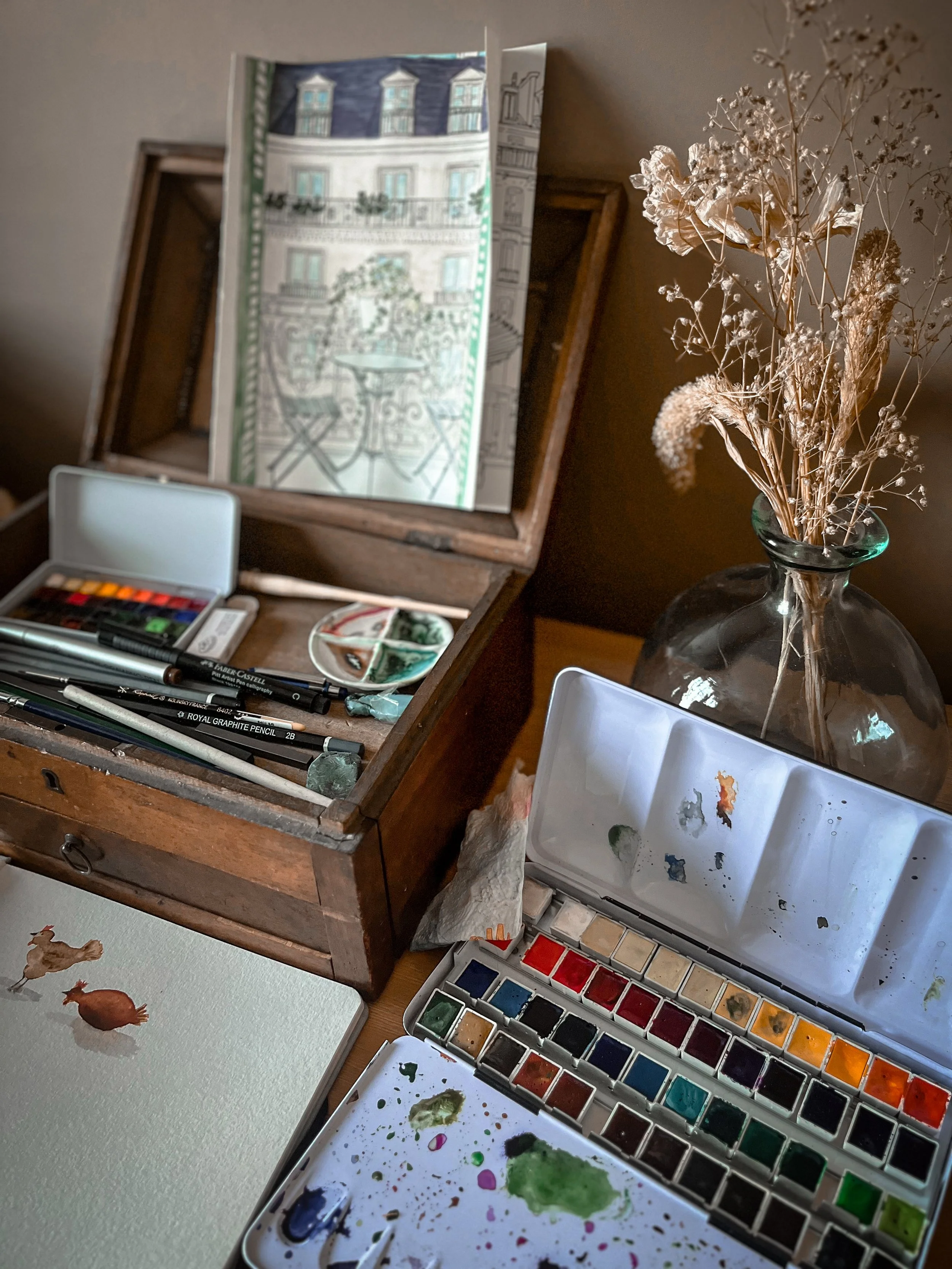 Une table de peinture avec des pinceaux, une palette, un ensemble de couleurs aquarelle, une boîte à peinture, un dessin d'un poulet, une vase avec des fleurs séchées, et un miroir posés sur un meuble en bois.