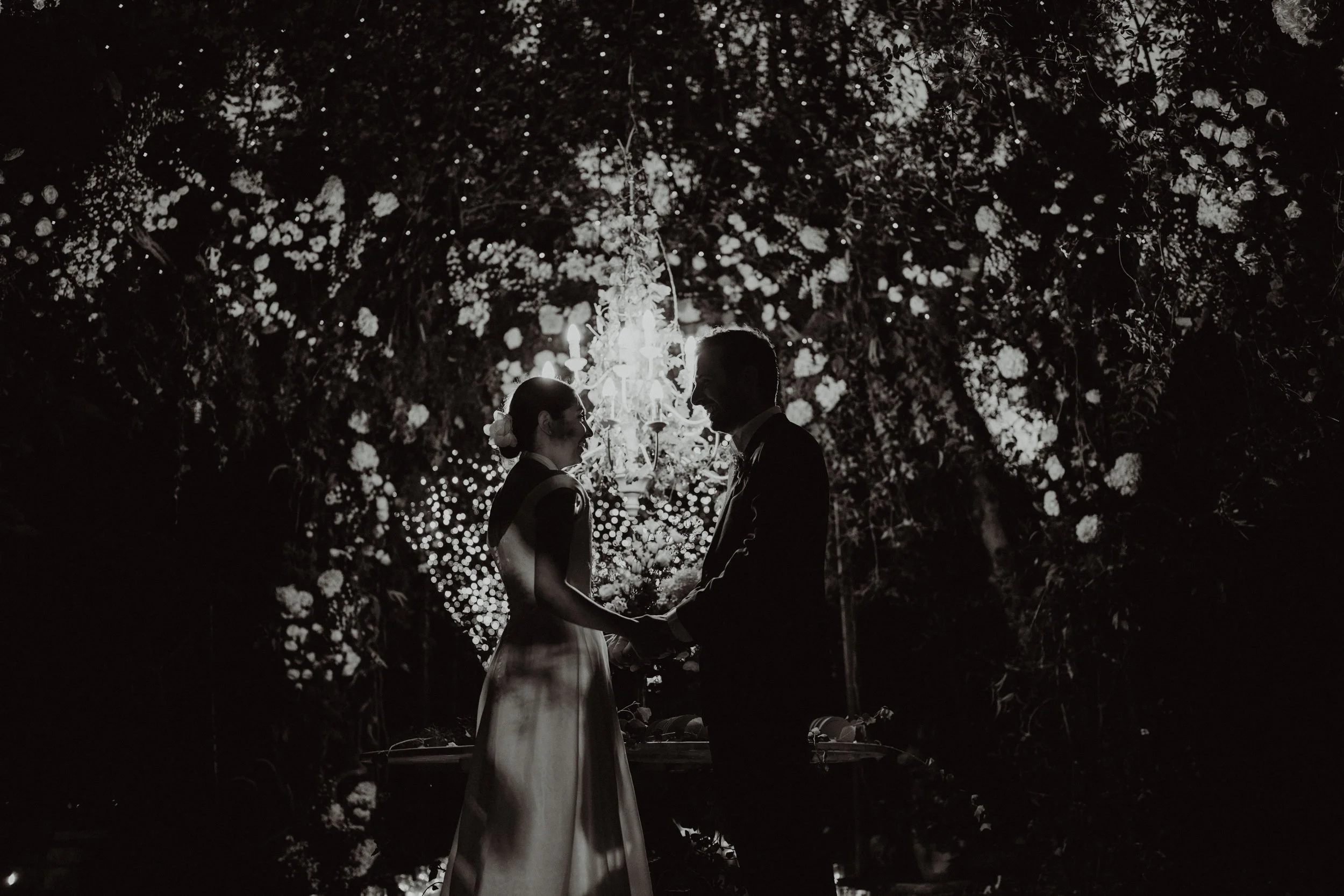 Silhouette di una coppia che si tiene per mano durante un matrimonio, circondata da un arco di fiori e luci soffuse, in un ambiente romantico di notte.