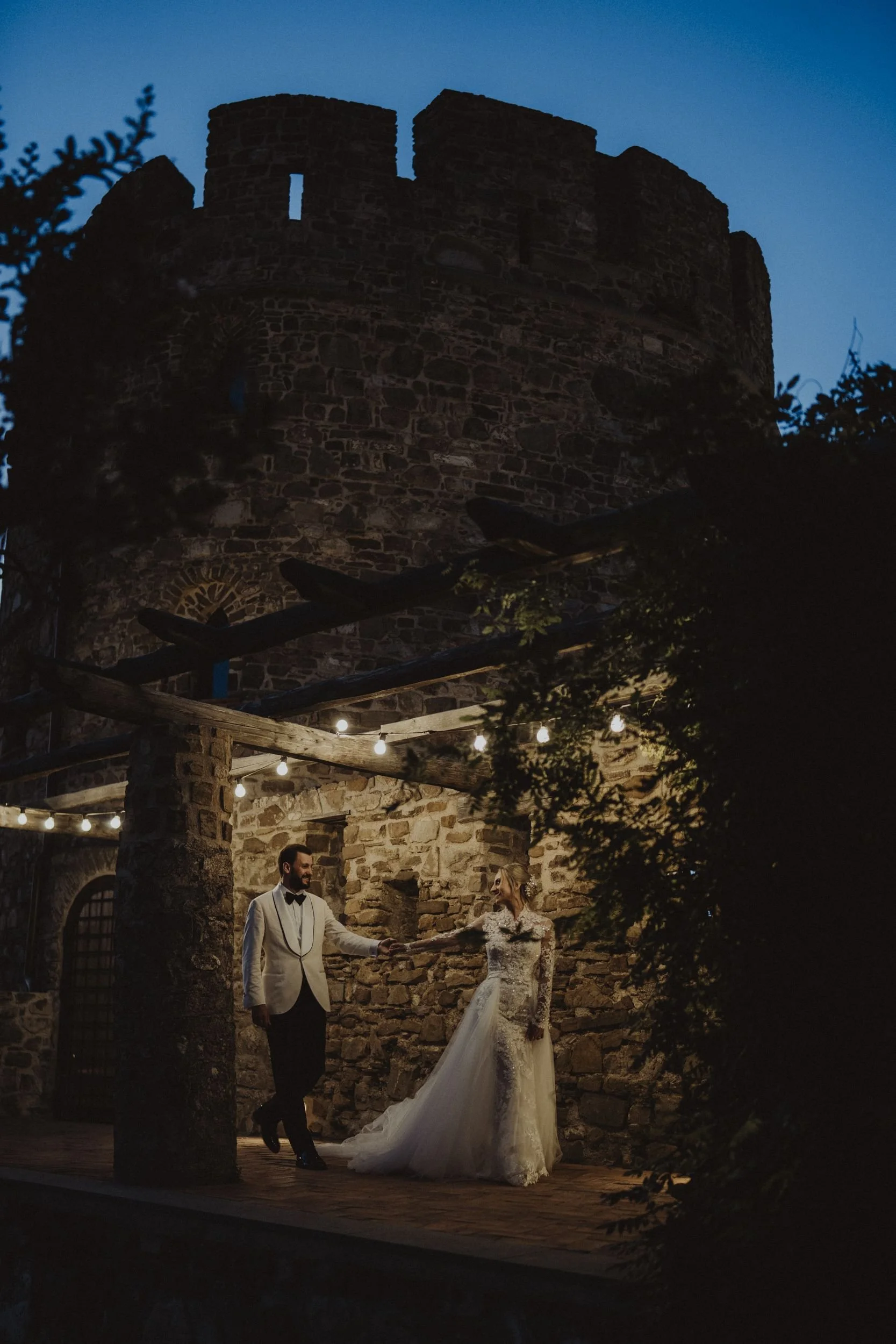 Una coppia di sposi in abiti da matrimonio balla sotto luci di stringa, con un'antica torre di pietra sullo sfondo in un cortile di pietra al tramonto.