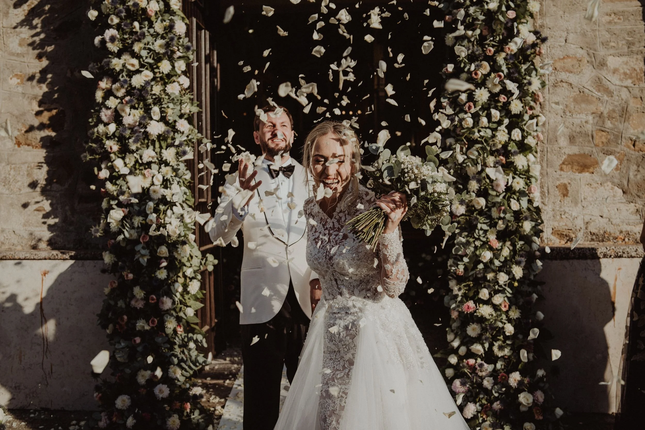 Una coppia di sposi felici che festeggiano il loro matrimonio sotto un arco decorato con fiori, mentre petali di fiori vengono lanciati nell'aria.