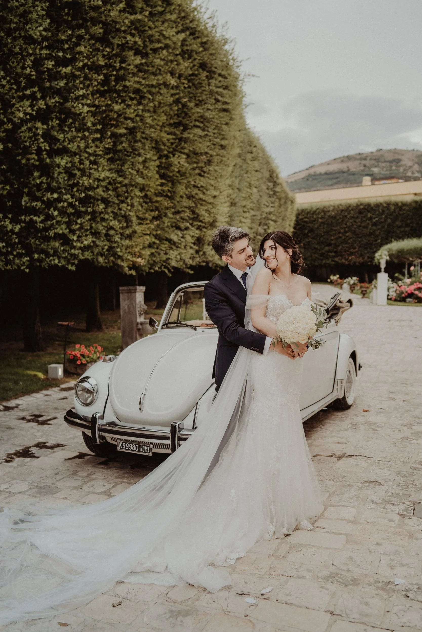 Una coppia di sposi in abiti da matrimonio si abbraccia davanti a una macchina d'epoca bianca, con un paesaggio di alberi e colline sullo sfondo.