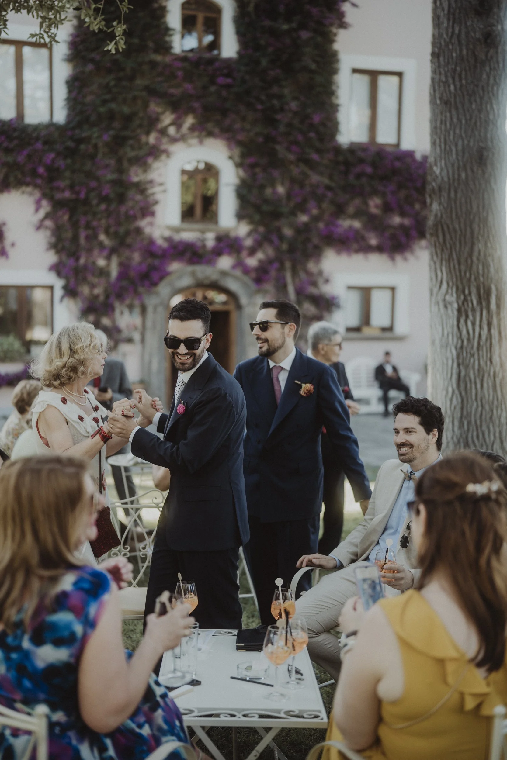 Un gruppo di persone che si divertono a un matrimonio all'aperto, con due uomini in abiti formali che ballano e una donna anziana che condivide un momento di gioia.