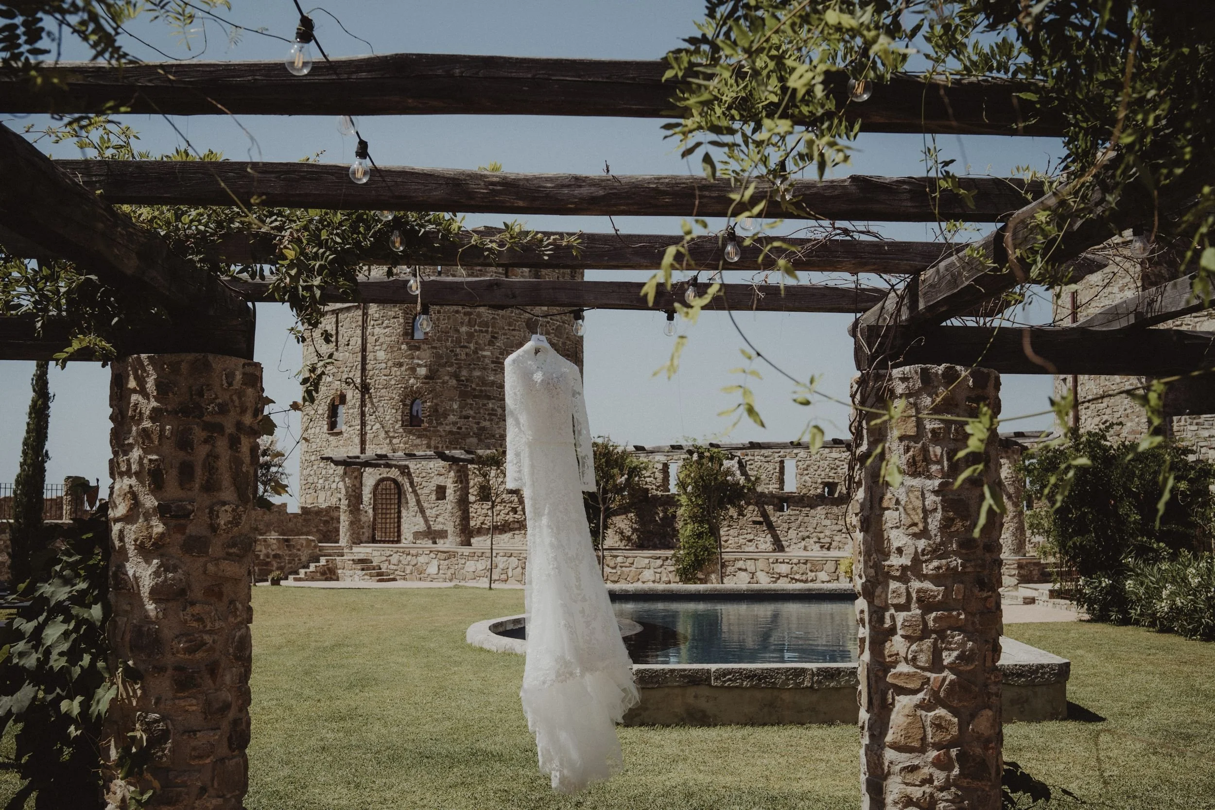 Vestito da sposa bianco appeso a un filo con luci di stringa, in un cortile con fontana e castello in pietra sullo sfondo.