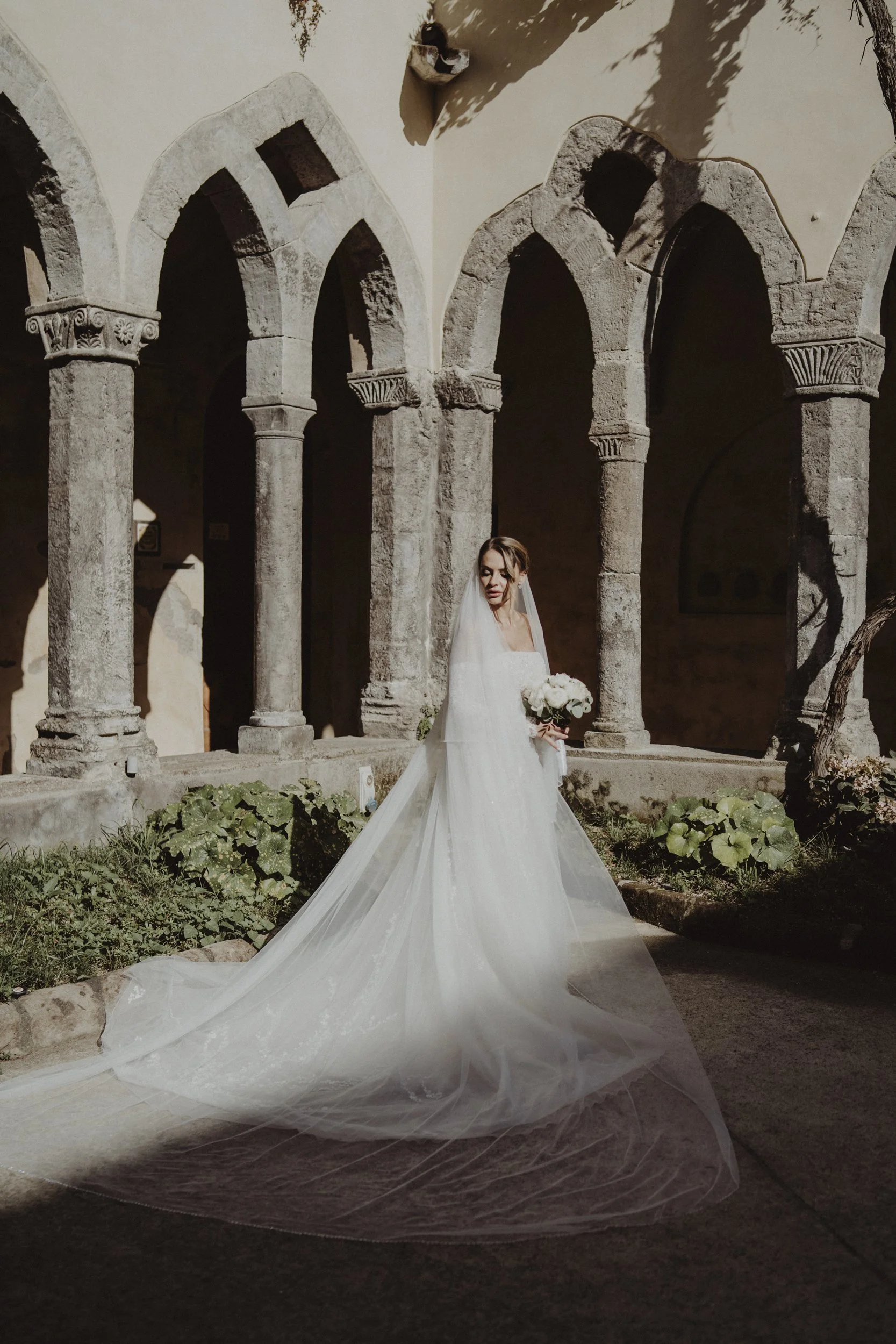 Sposa in abito bianco con velo lungo, che tiene un bouquet di fiori, in un cortile con archi antichi in pietra.
