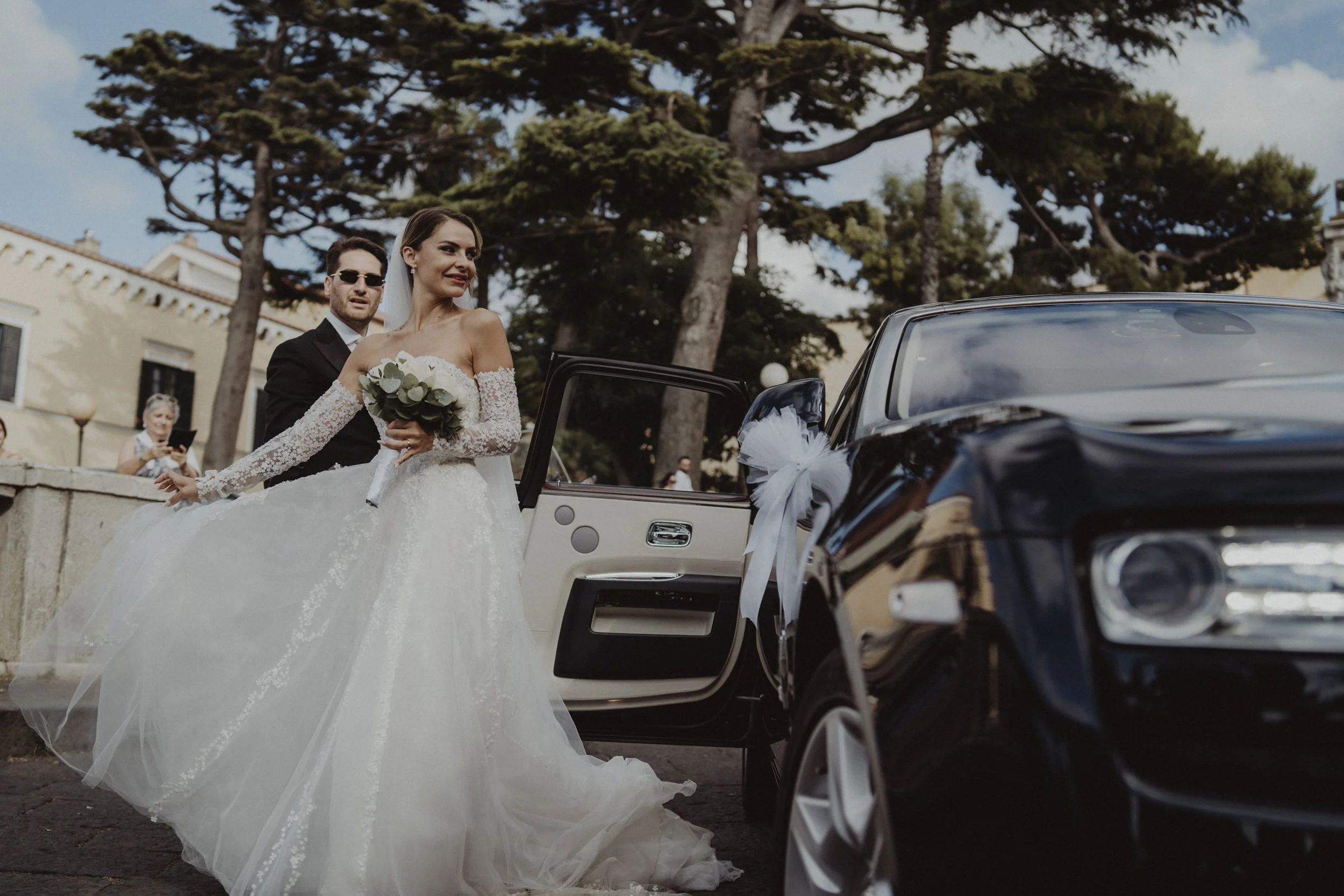 Una coppia di sposi lascia un matrimonio con una macchina nera decorata con un fiocco bianco, la sposa tiene un bouquet di fiori e indossa un abito da sposa lungo e in pizzo, mentre il marito indossa un abito scuro. Sullo sfondo ci sono alberi e pers