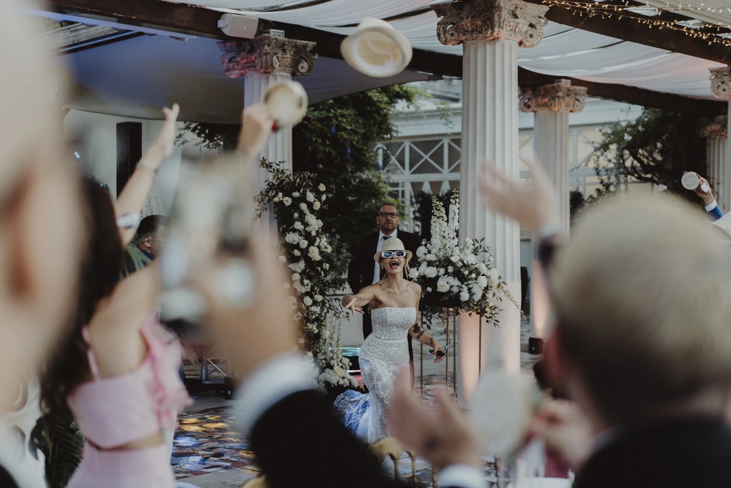 Una donna in abito da sposa che balla e ride, circondata da amici e parenti in un matrimonio all'aperto con decorazioni floreali e colonne classiche.