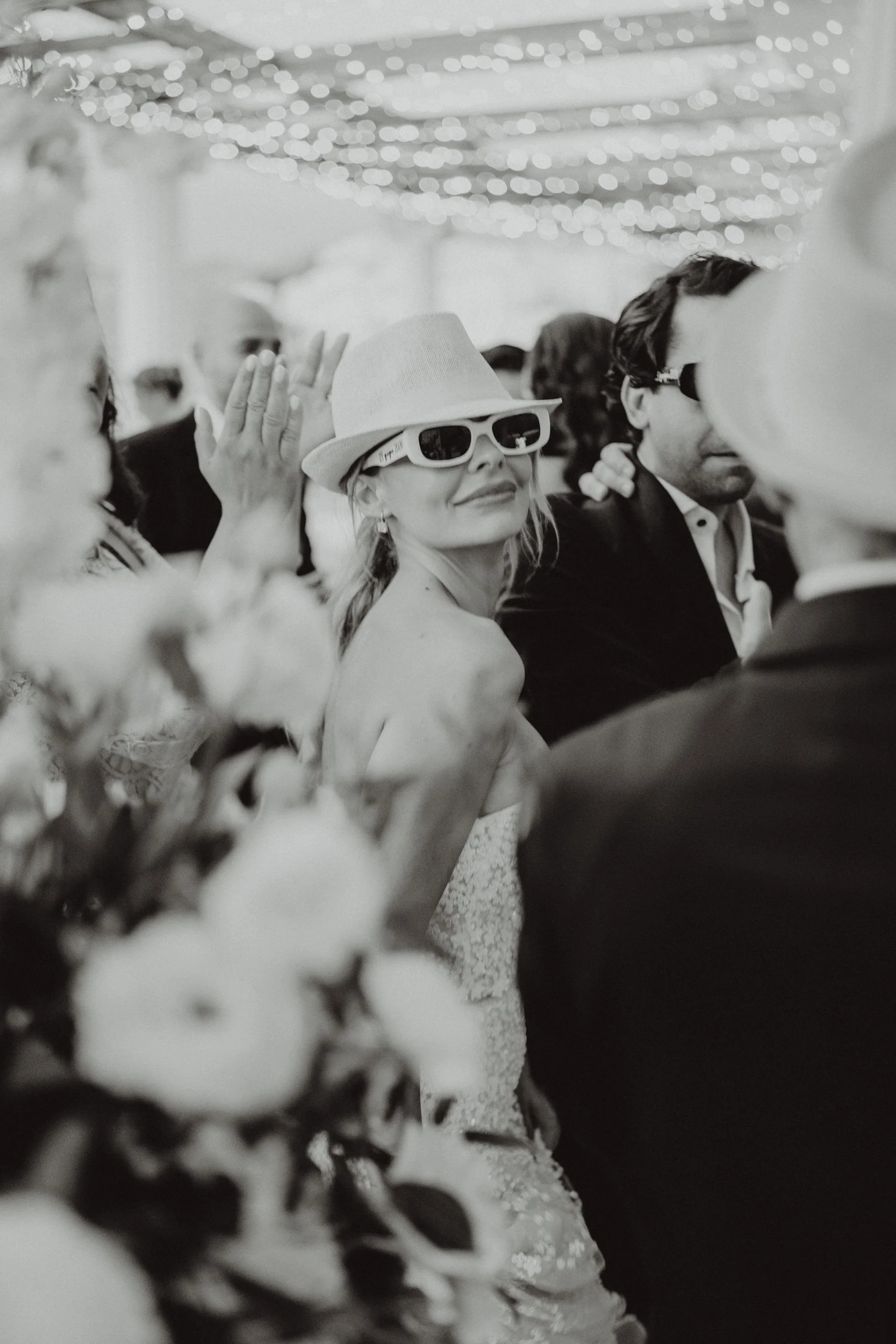 Donna con cappello e occhiali da sole in un evento sociale, sorridente, tra persone eleganti.