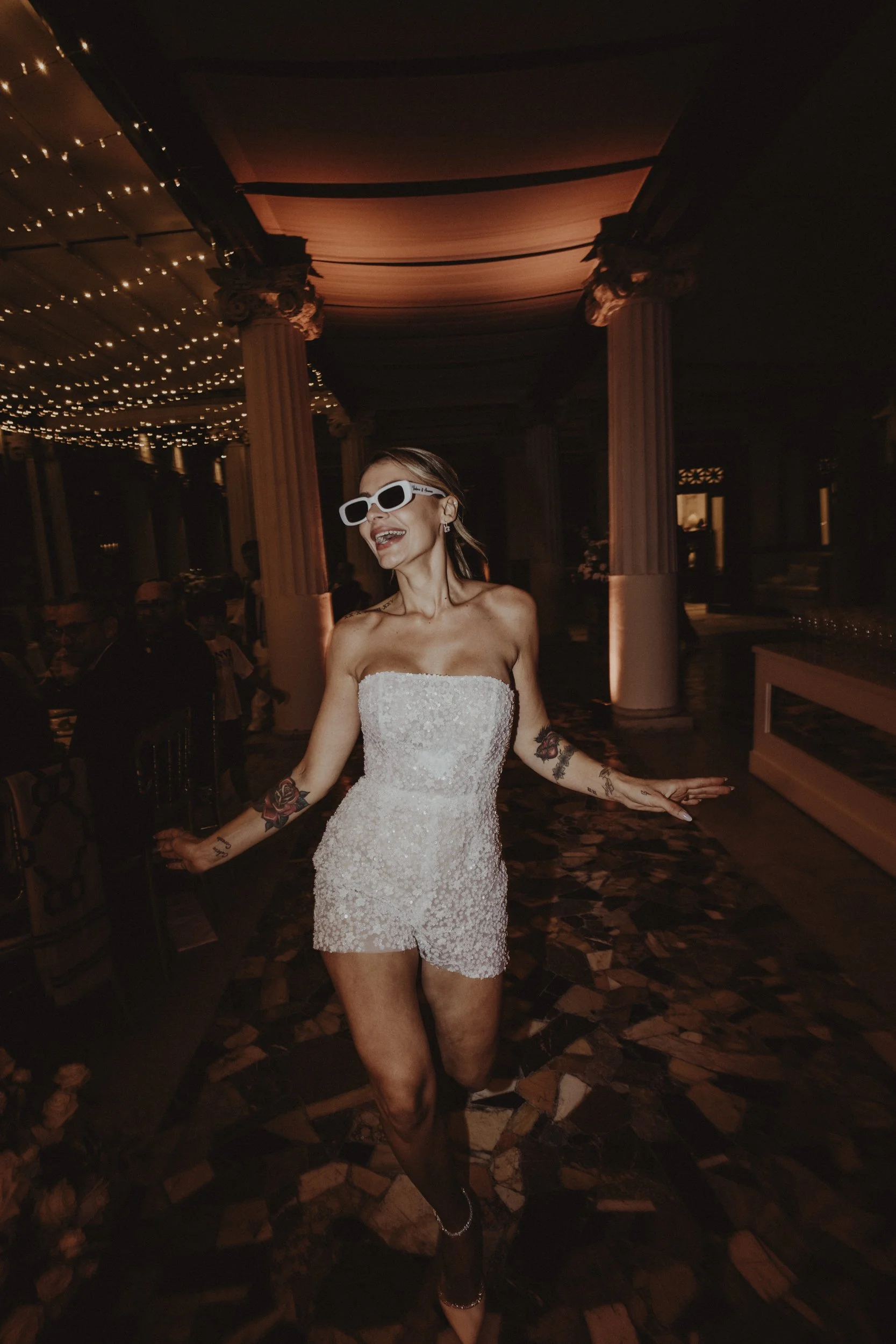 Una donna con vestito bianco, occhiali da sole bianchi, tatuaggi e sorriso balla in un locale elegante decorato con luci soffuse e colonne.